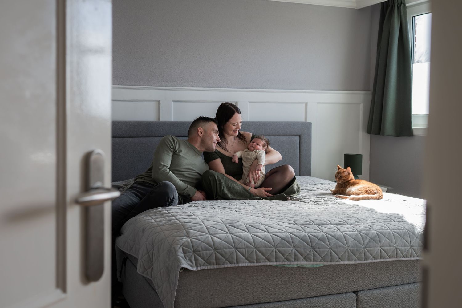 Couple relaxing on a gray upholstered bed with a small dog, in a cozy modern bedroom with green curtains.