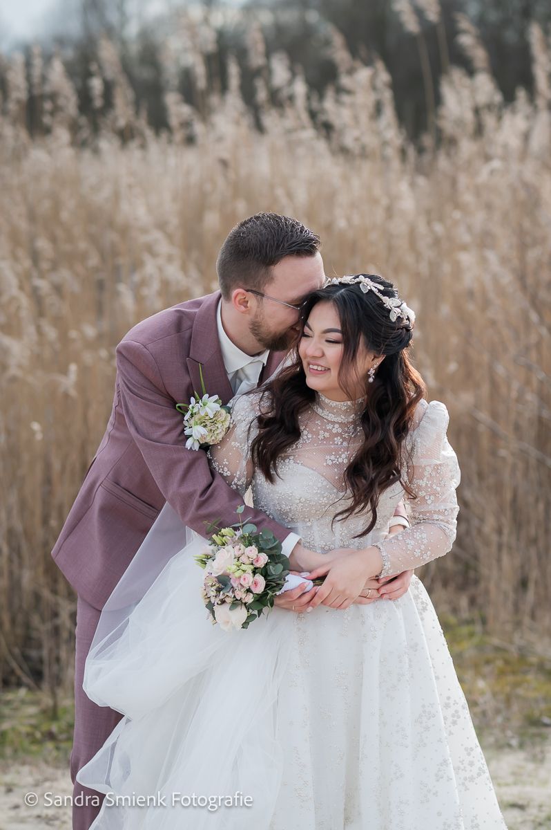 Groom in pink suit kissing bride in lace wedding dress outdoors near tall reeds, holding a bouquet.