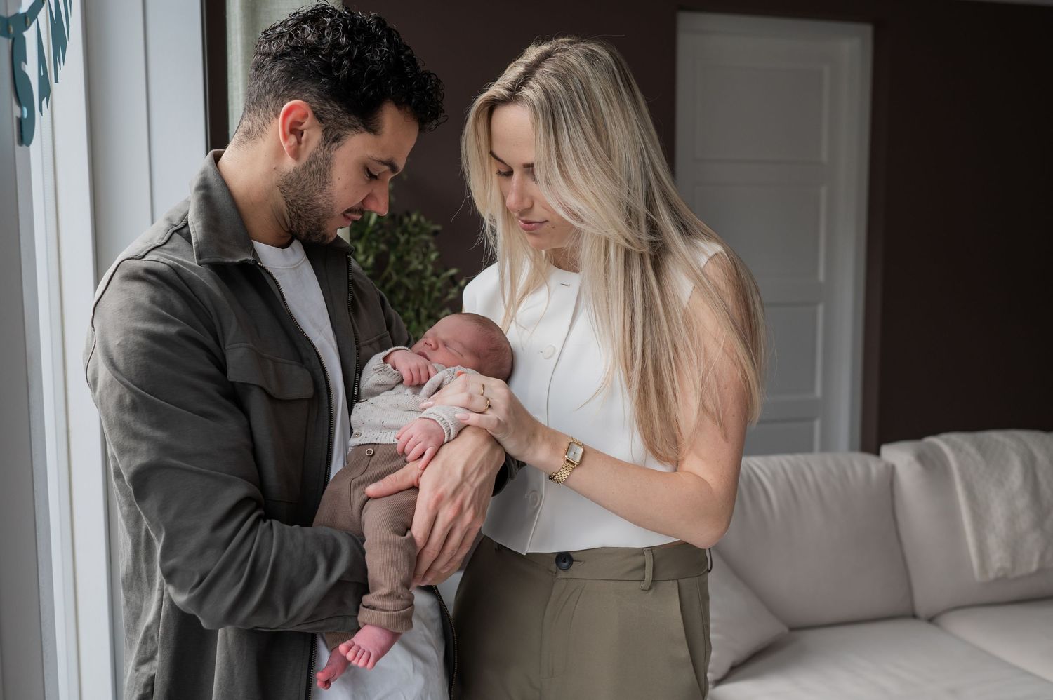 New parents standing together and looking down at their newborn baby in a home portrait.
