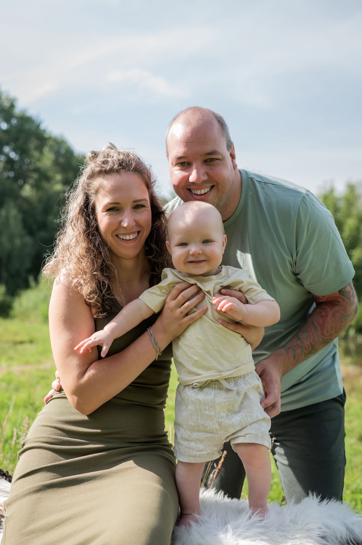 Happy family portrait outdoors with smiling parents holding their baby on a sunny day in a green field.