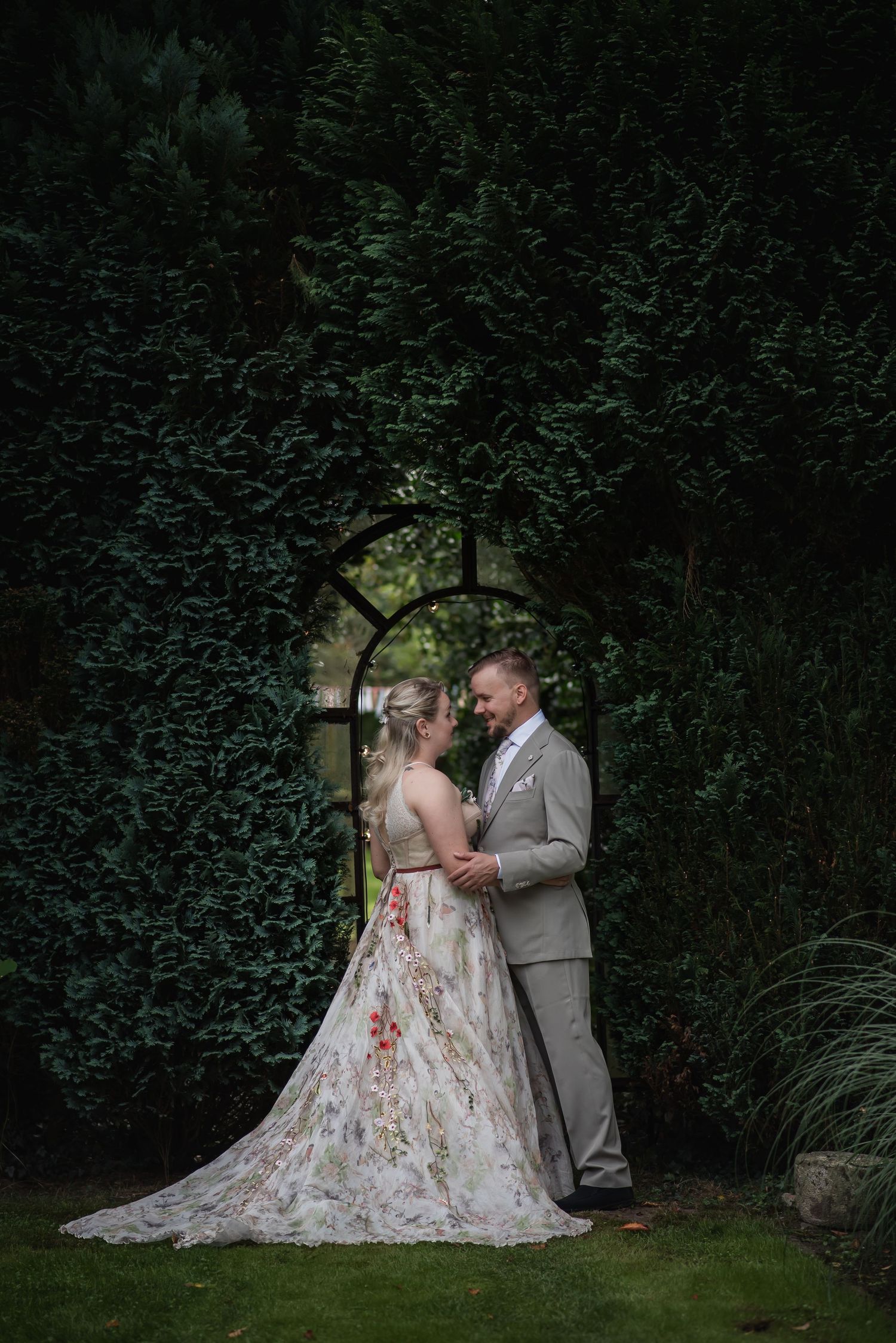 Bride in floral gown and groom in gray suit embrace before lush green hedge with arched mirror at garden wedding.