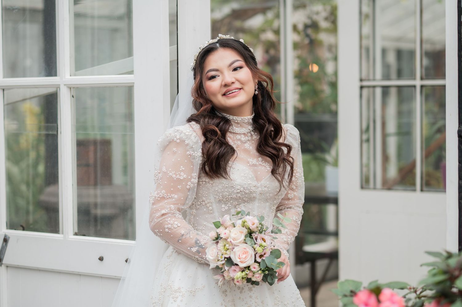 Smiling bride in lace long-sleeve wedding gown holding pink bouquet, standing by white windows.