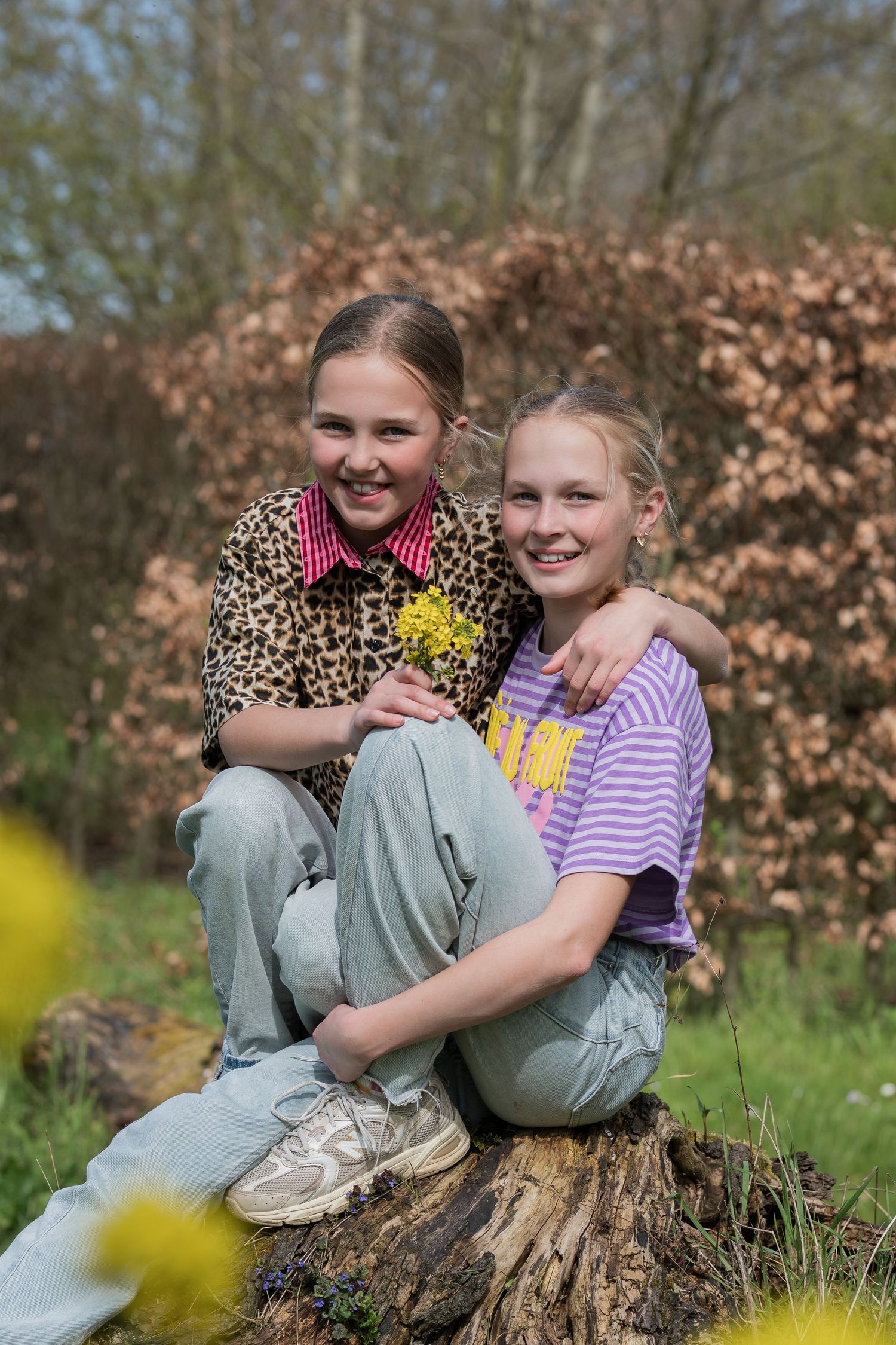 Two smiling girls sitting on a tree stump outdoors, one in leopard print and one in a purple striped shirt.