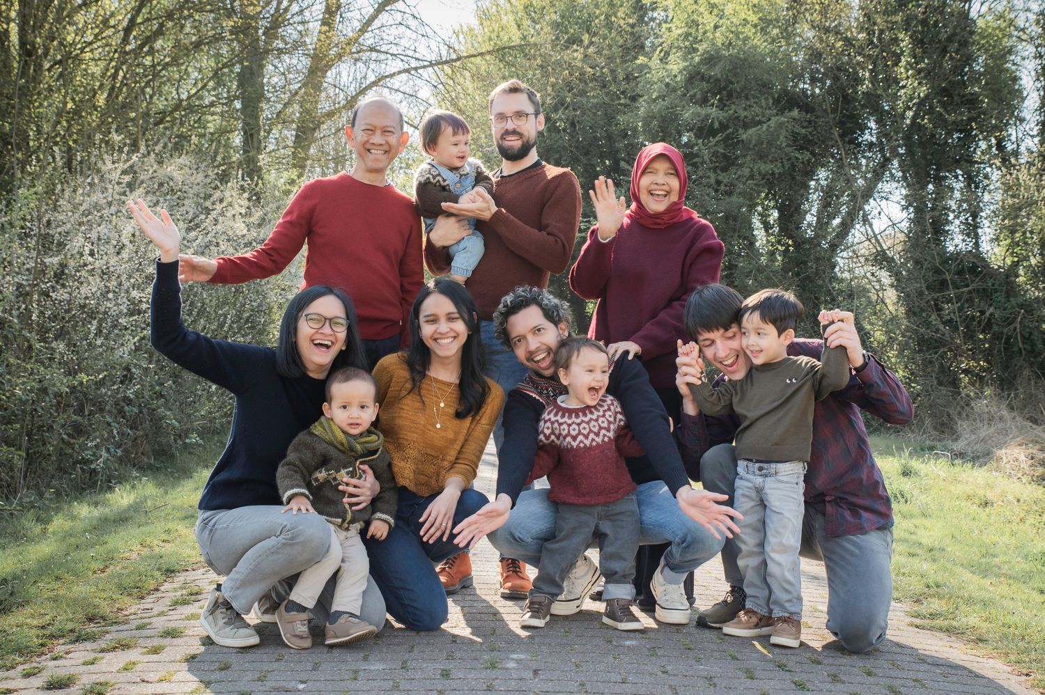 Large multigenerational family group smiling and waving together outdoors in a park setting on a sunny day.