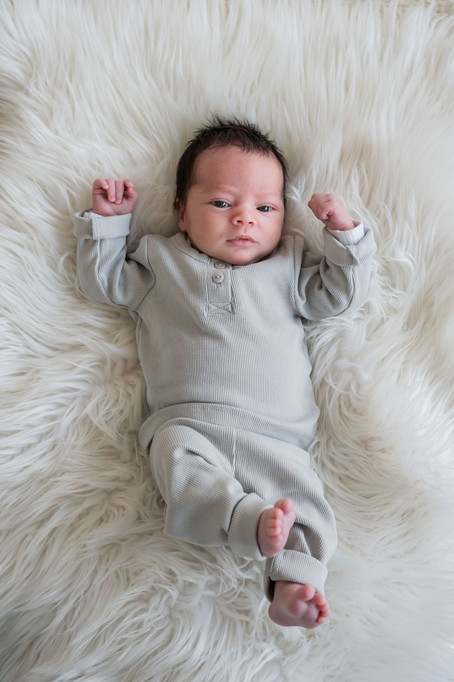 Alert newborn baby lying on a fluffy white rug with arms raised in a grey outfit.