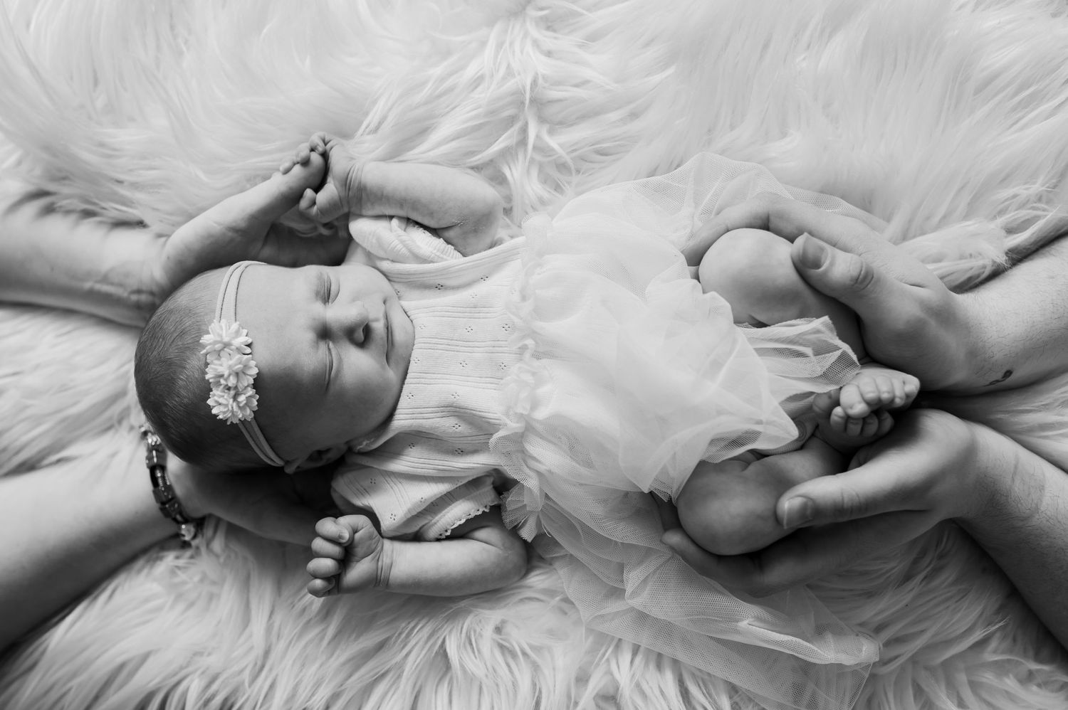 Black and white newborn photo of a baby girl in white dress and floral headband, cradled by adult hands on fluffy fur.