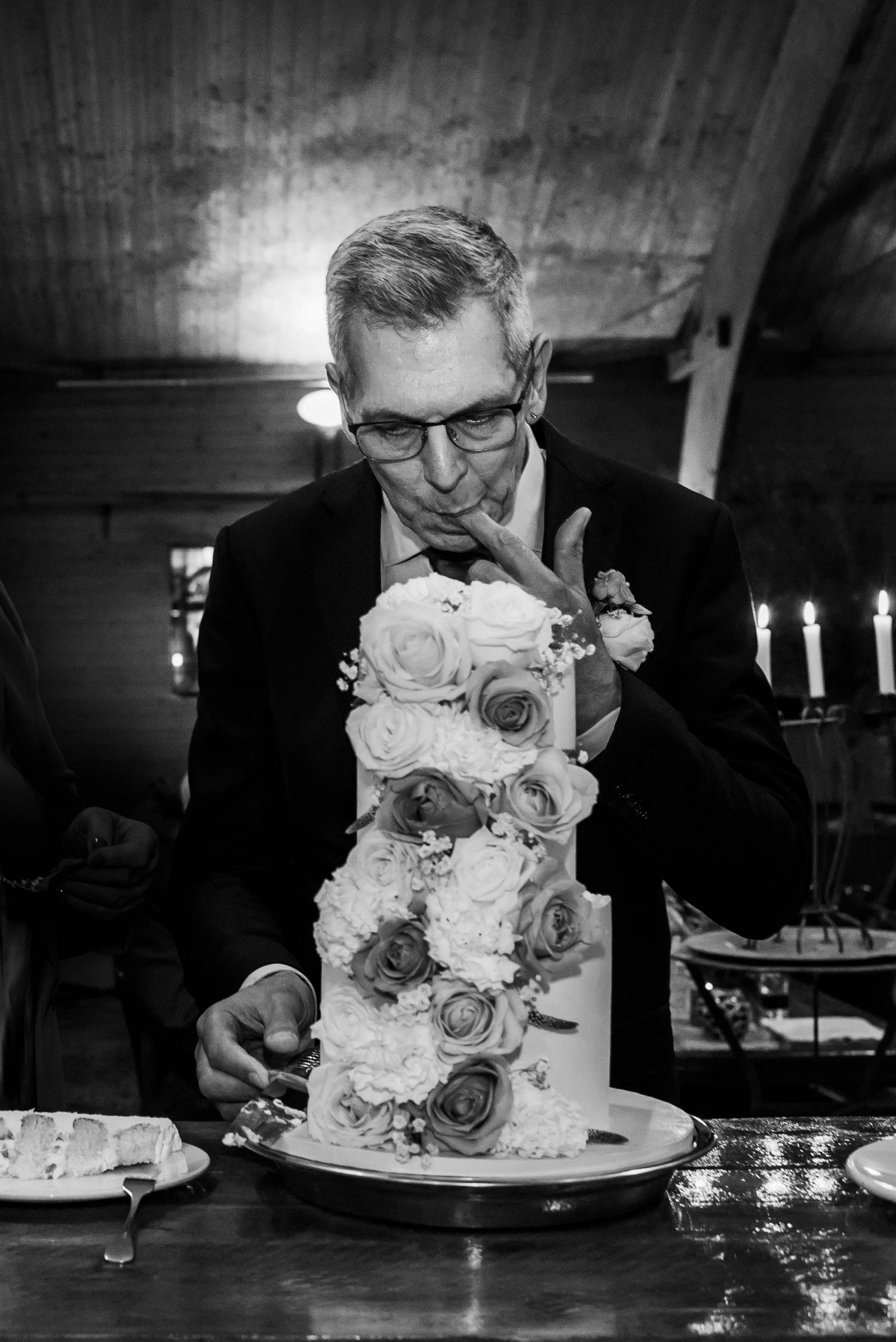 Black and white photo of a man in a suit tasting wedding cake beside a floral tiered cake at a reception.