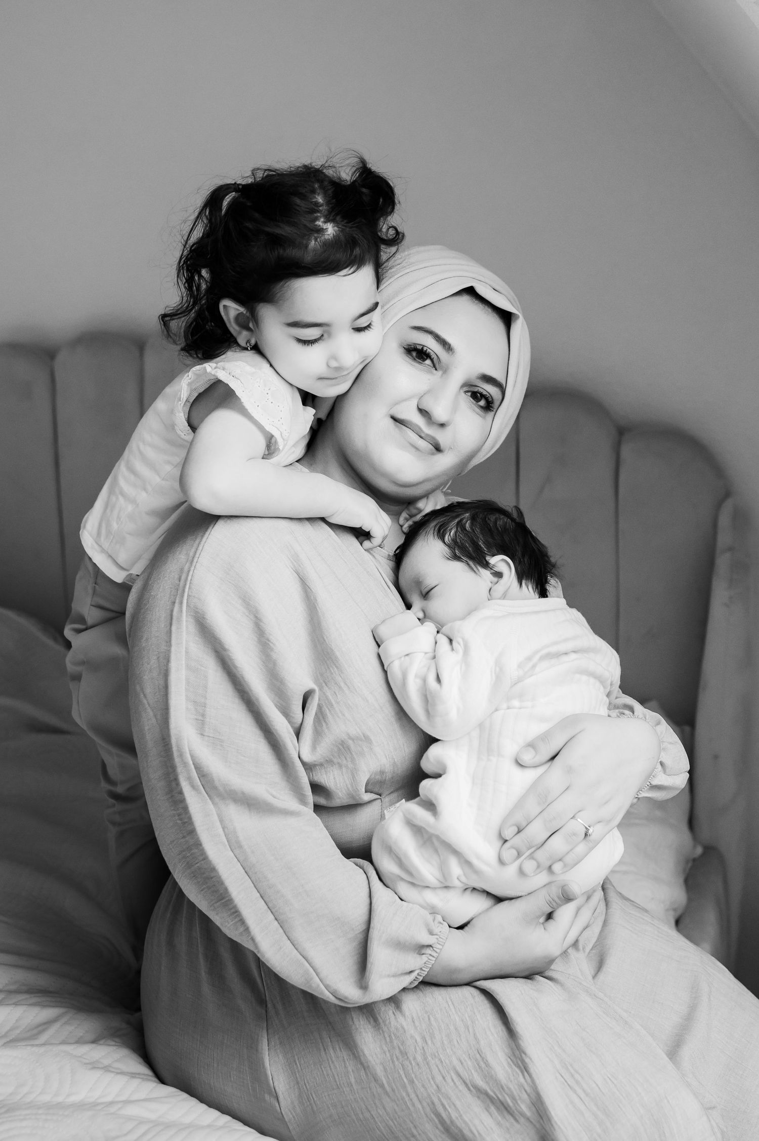Black and white photo of a mother in hijab tenderly holding a newborn and toddler while sitting on a bed. Lelystad, Almere, Flevoland