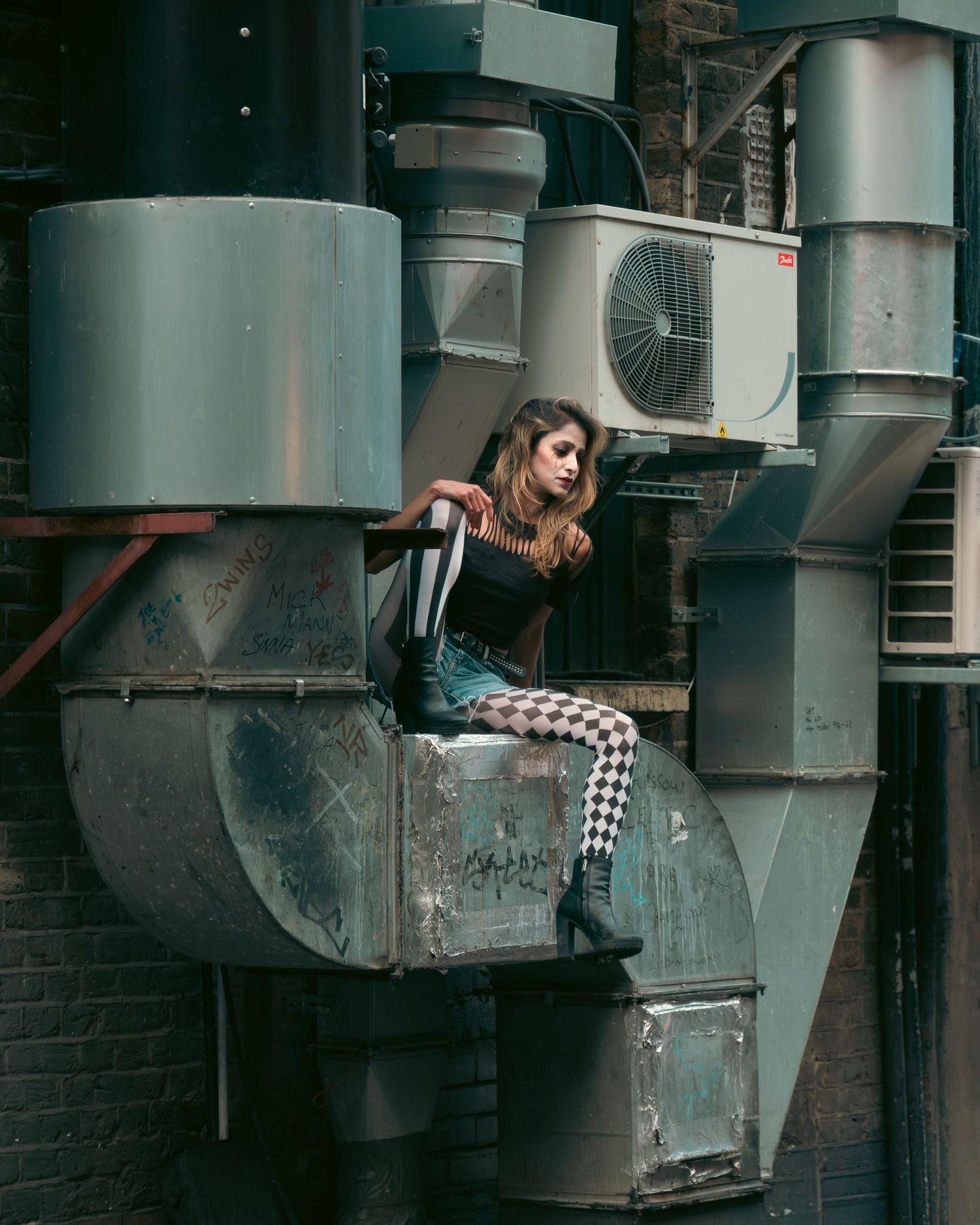 Donna con calze a scacchi seduta su condotti industriali metallici. / Woman in checkered tights sitting on industrial metal ducts. ME visual