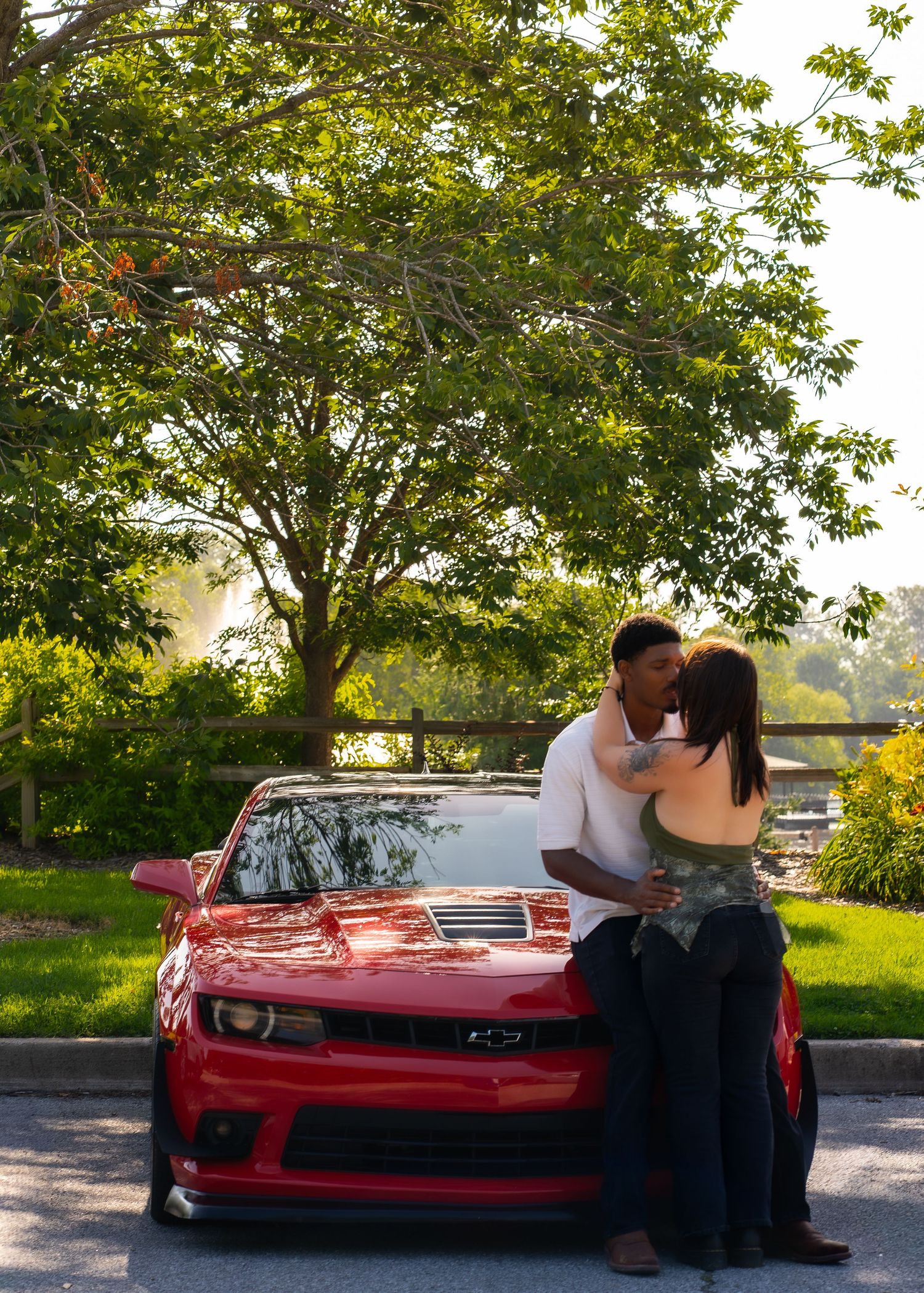 Couple sharing a romantic kiss leaning against a red Chevrolet Camaro outdoors with lush green trees in background.