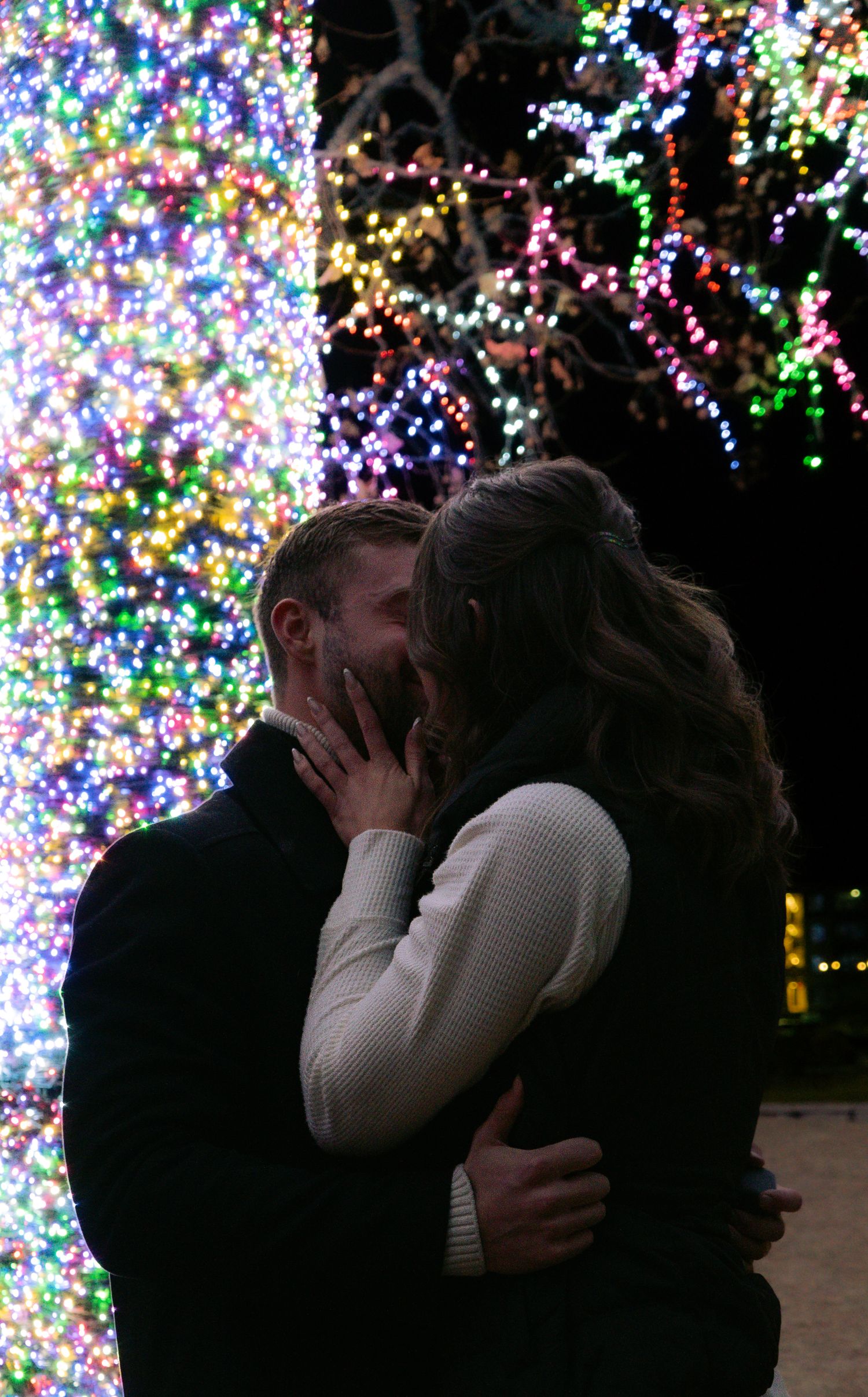 Couple sharing a romantic kiss surrounded by colorful Christmas lights on trees at night.