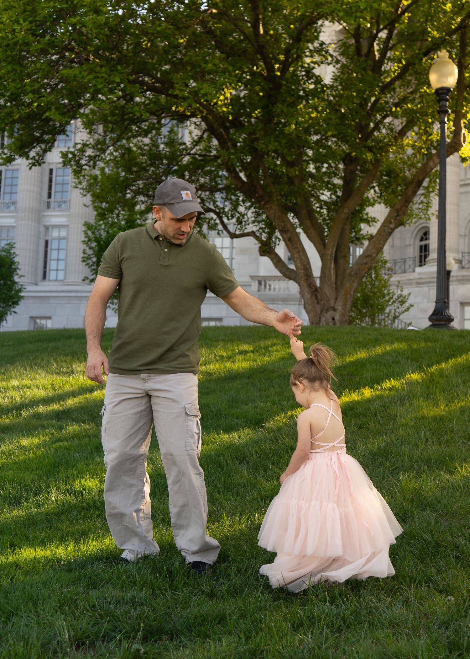 Father tenderly spinning his little girl in a pink tulle dress on a green lawn at golden hour at the Missouri Capitol. Shelby Renee Studios