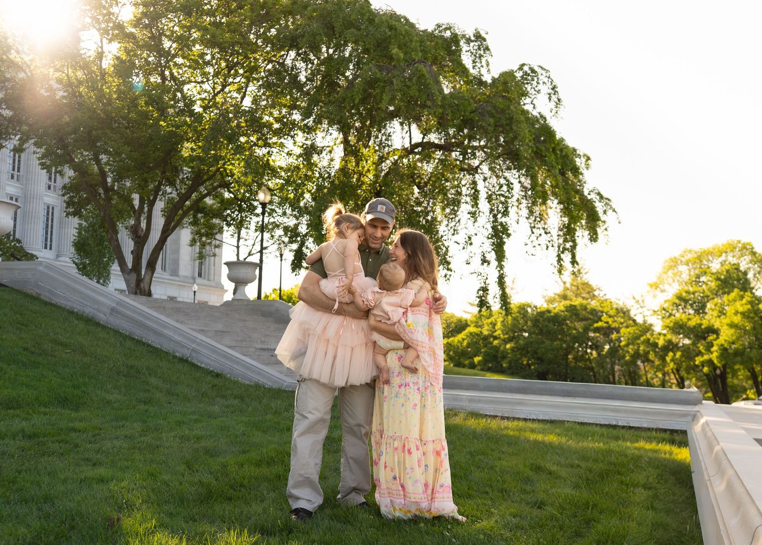 Happy family embracing on a sunlit green lawn at the Missouri Capitol with a white mansion and weeping willow in background. Shelby Renee