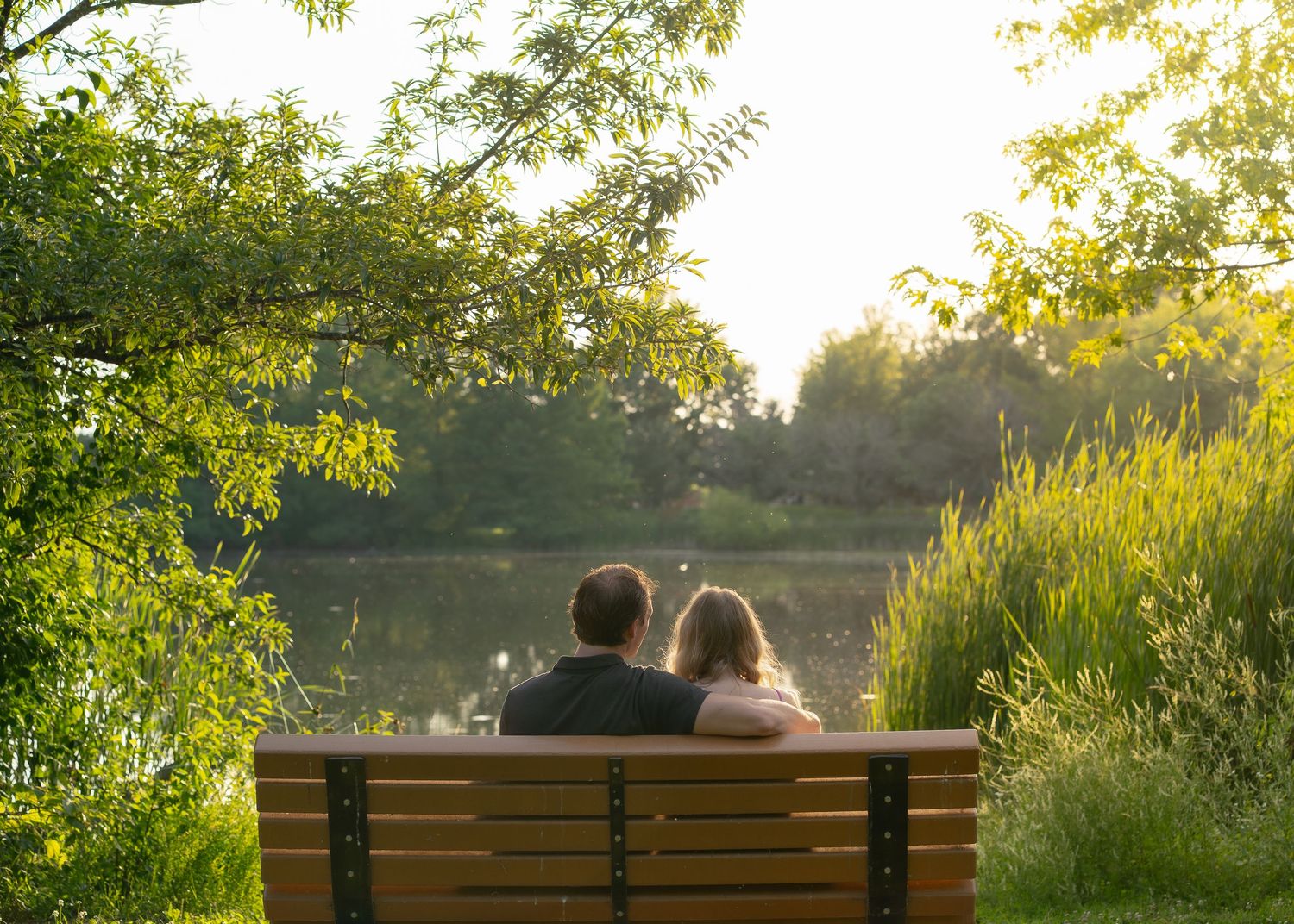 Couple sitting closely on a bench overlooking a peaceful sunlit lake surrounded by trees. Bethel Park Columbia MO Shelby Renee Studios