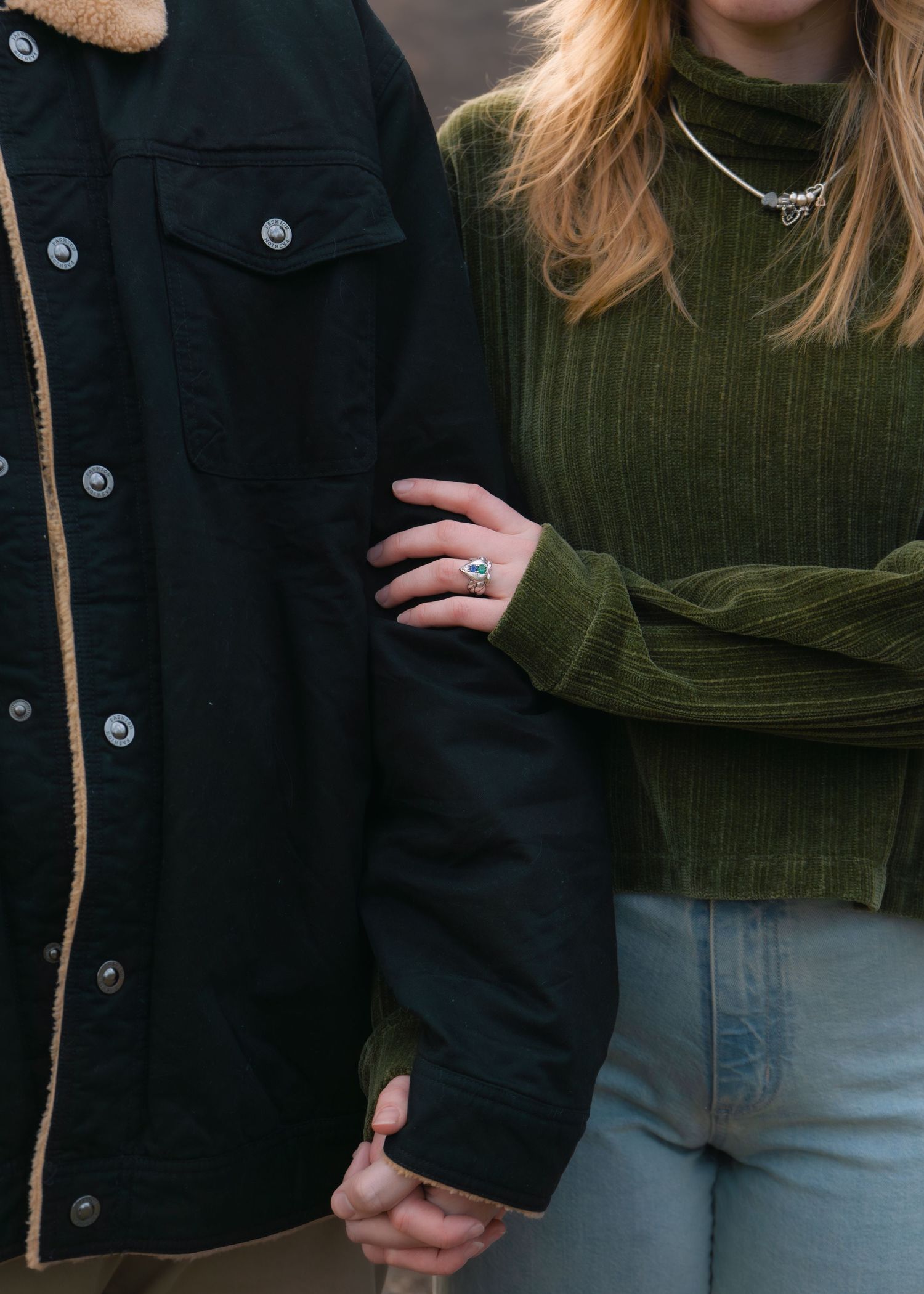 Couple holding hands, woman in green ribbed turtleneck with ring, man in black sherpa jacket, casual outdoor setting.
