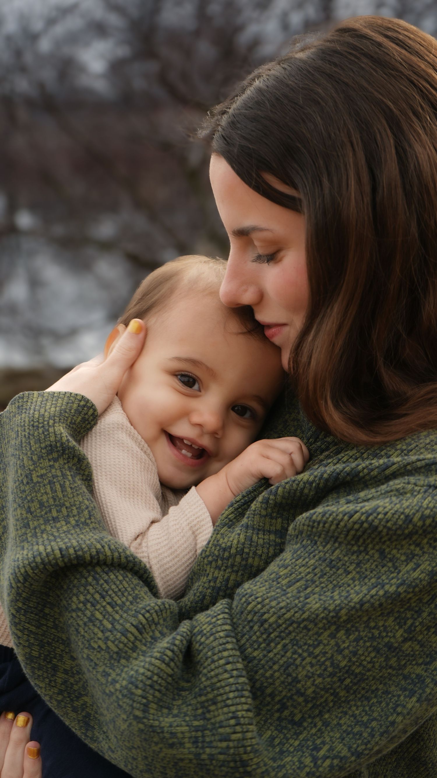A mother with brown hair in a green sweater tenderly kisses her smiling toddler outdoors in winter.
