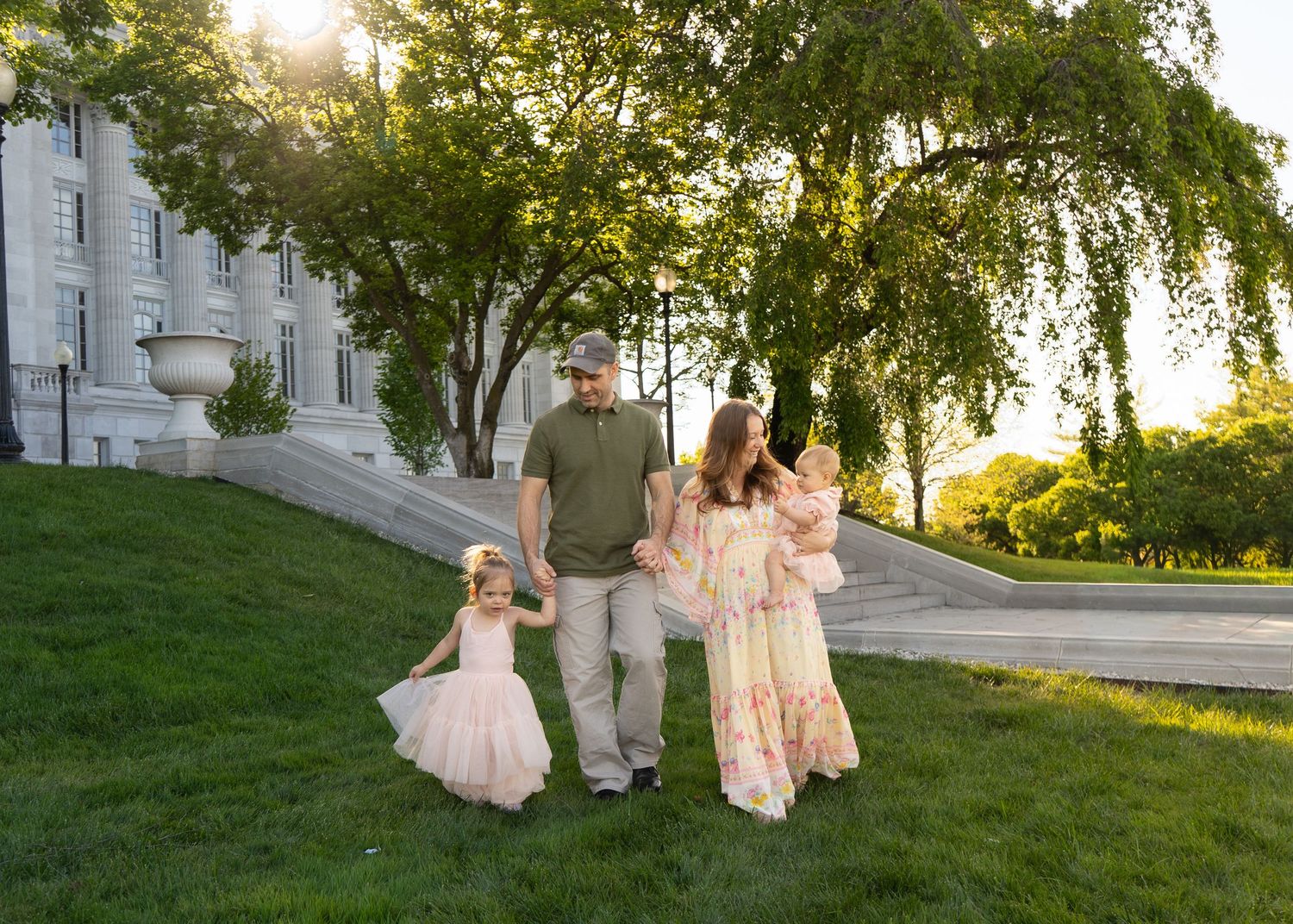 Family of four walking on green grass near historic building during golden hour, parents holding children in pink dresses.