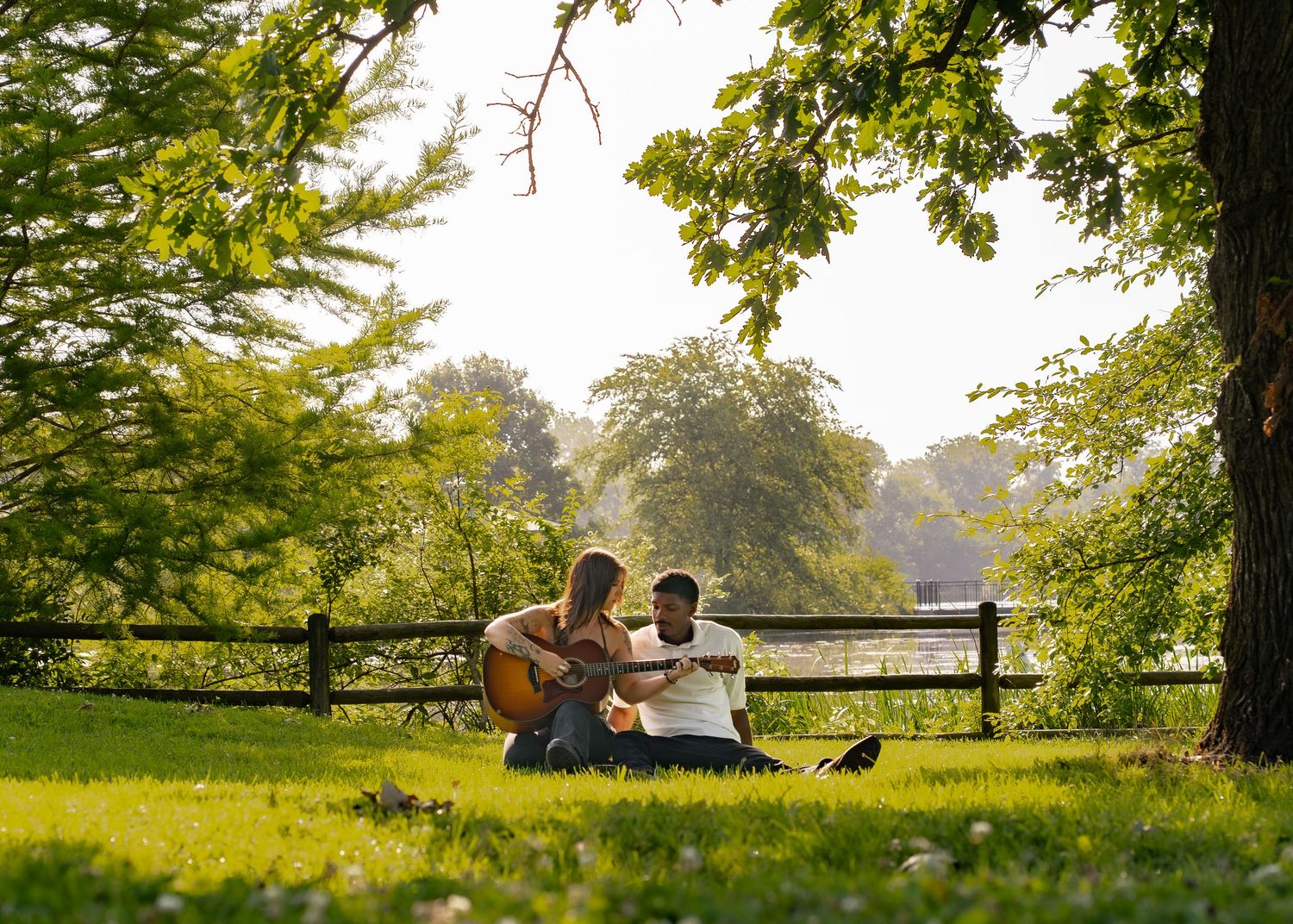 Couple sitting on lush green grass under tree by fence as man plays acoustic guitar in sunny park. Stevens Lake Columbia MO Shelby Renee