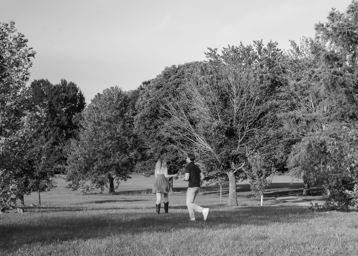 Black and white photo of a couple holding hands walking through a lush green park surrounded by tall trees.