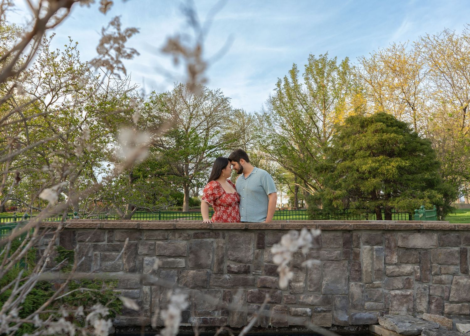 A couple shares a romantic moment atop a stone wall surrounded by spring blossoms and lush green trees in a park.