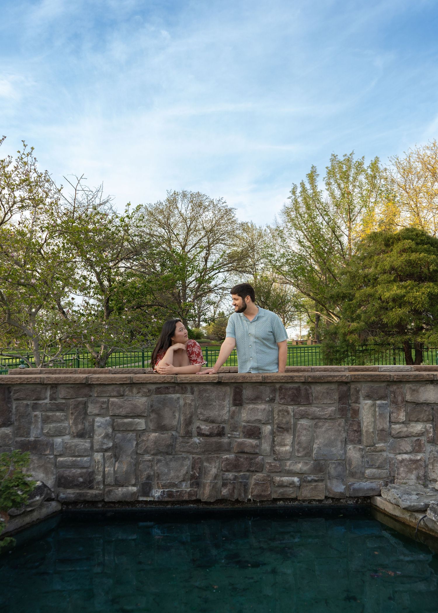 Young couple gazing at each other beside a stone wall pool with blue sky and trees behind them. Shelter Garden Columbia Shelby Renee Studios