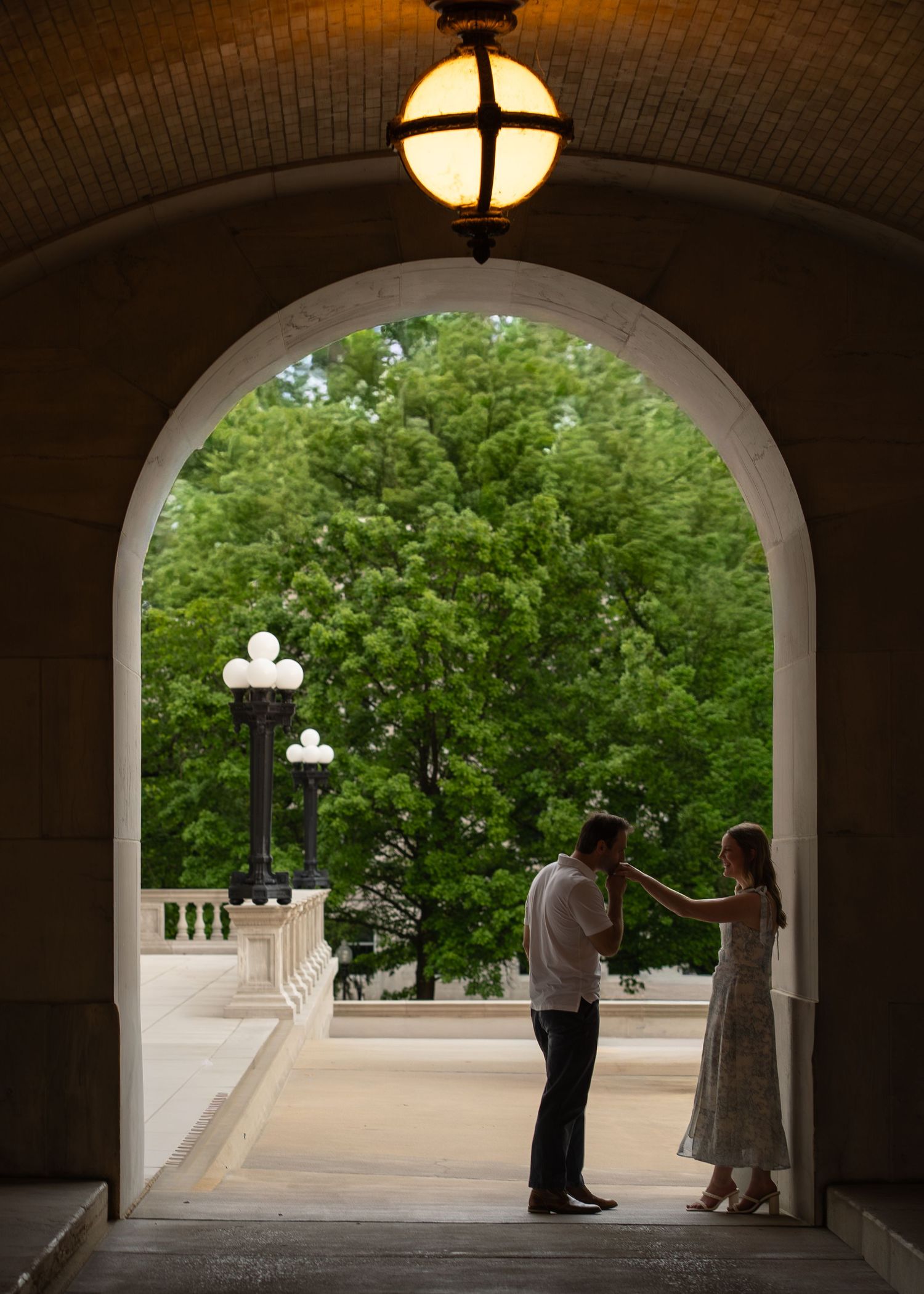Couple sharing a romantic moment framed by an elegant stone archway with glowing lantern and lush green trees. Missouri Capitol Shelby Renee