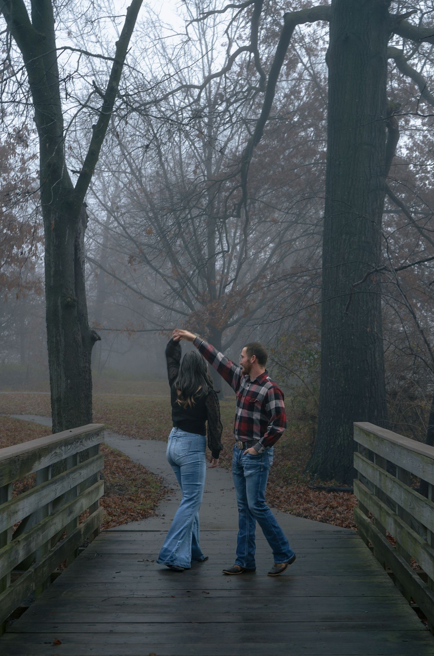 Couple dancing on a wooden bridge in a foggy autumn park surrounded by bare trees and fallen leaves.