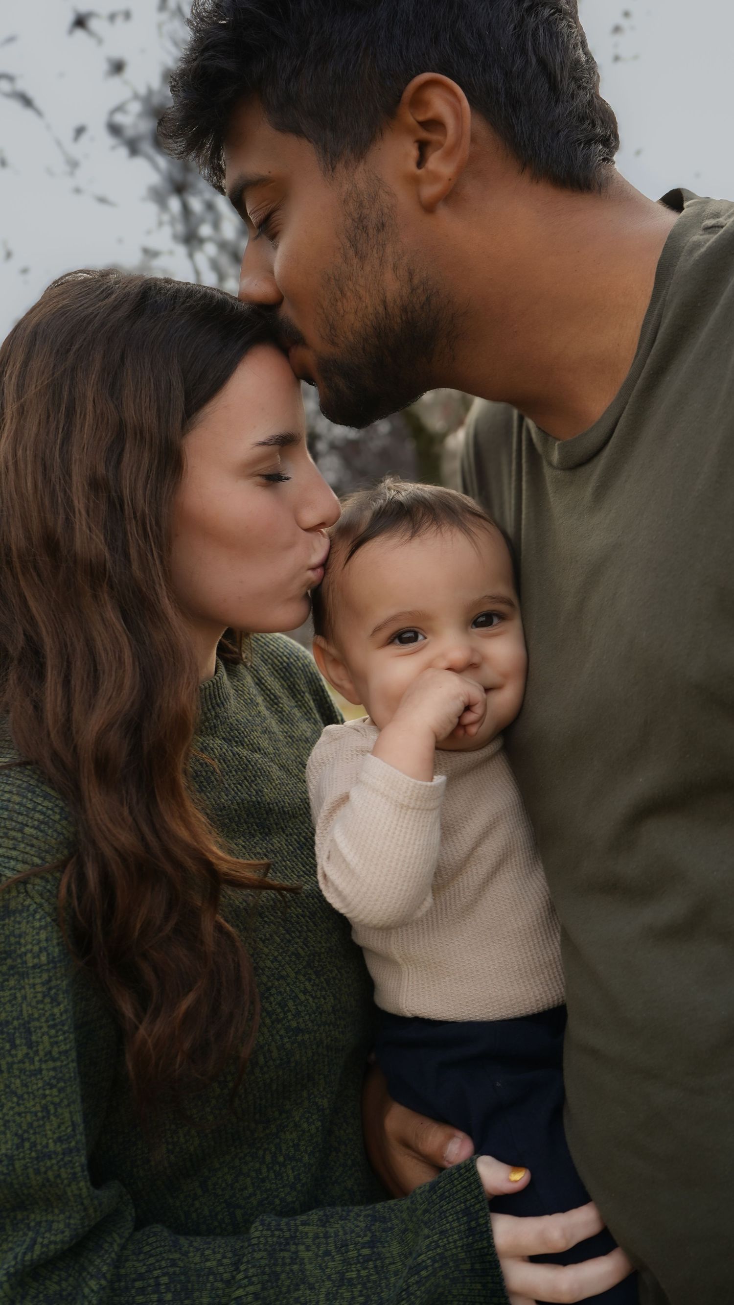 A young couple kisses their adorable baby on the cheeks in a tender family portrait outdoors.