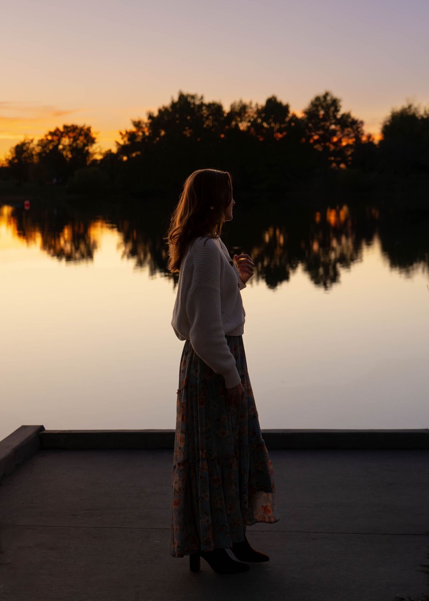 Woman in floral skirt holding wine glass on dock at sunset with lake reflection and silhouetted trees.