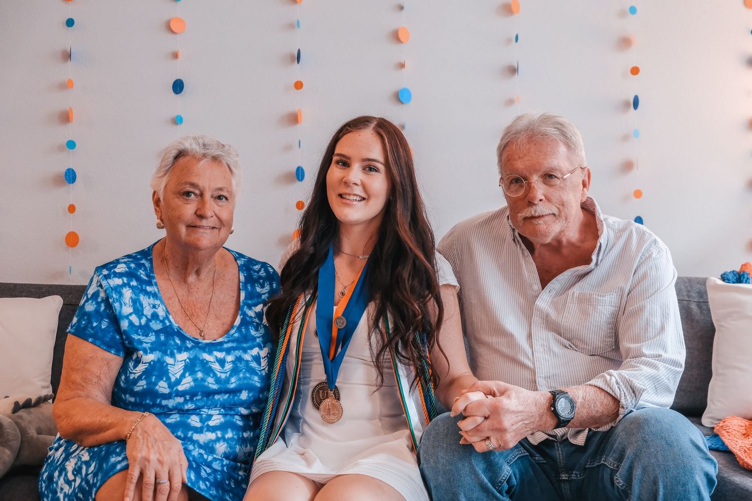 Young woman wearing a bronze medal posing with smiling grandparents on a couch at a celebration.