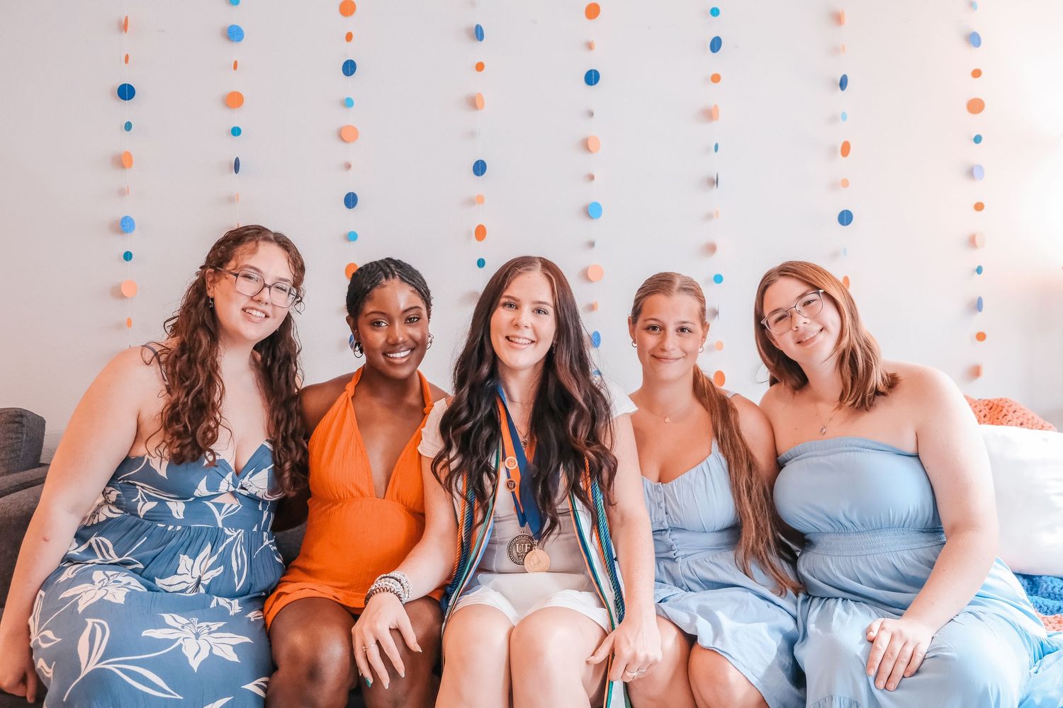 Five smiling women in colorful dresses sit together on a couch with festive circle garland decorations behind them.