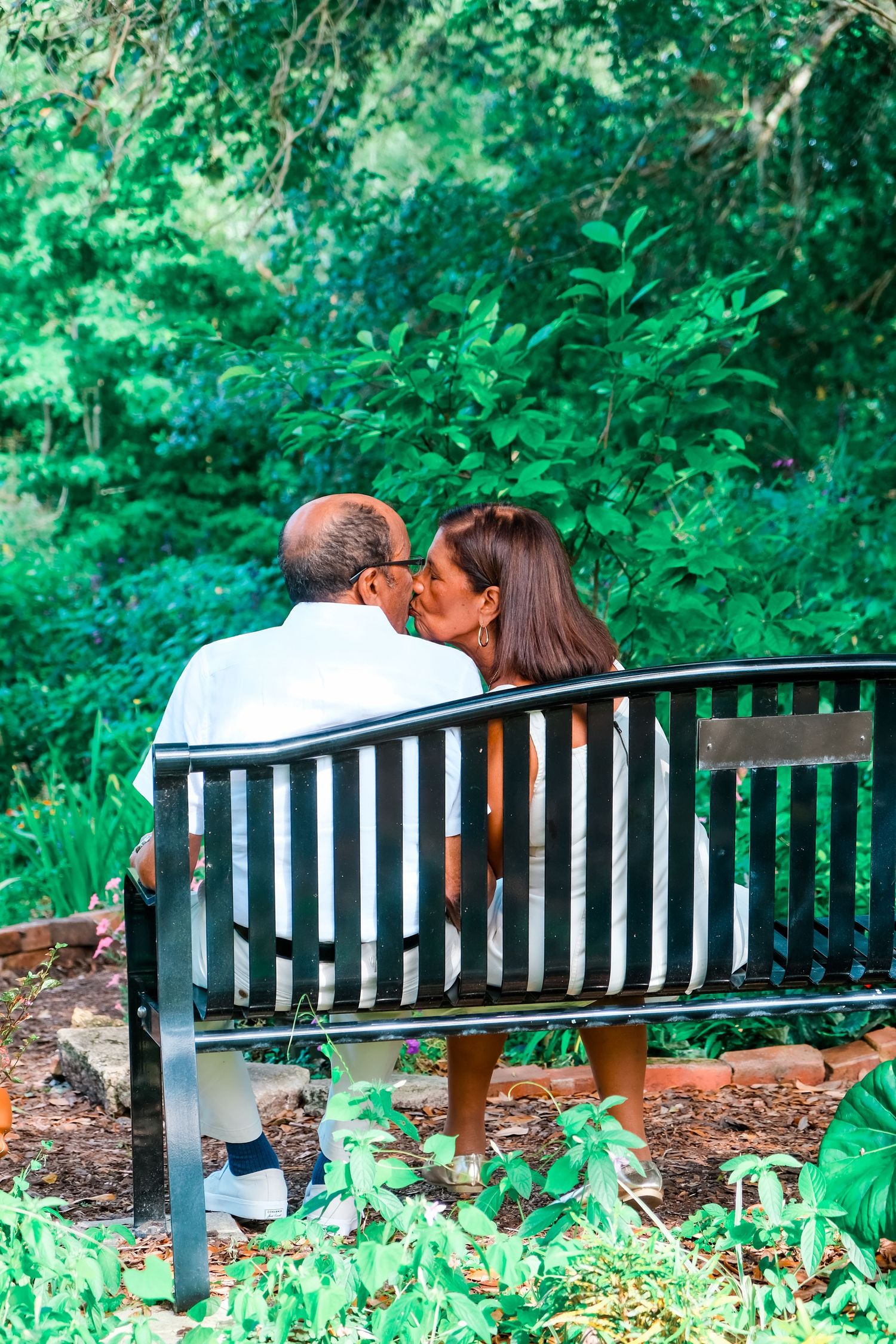 Couple sharing a romantic kiss on a park bench surrounded by lush green trees and foliage.