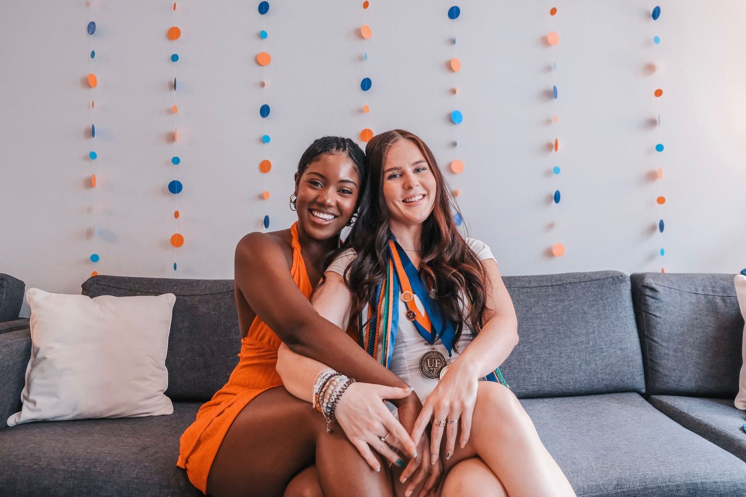 Two smiling women sitting on a gray couch in front of colorful circle garland decorations.