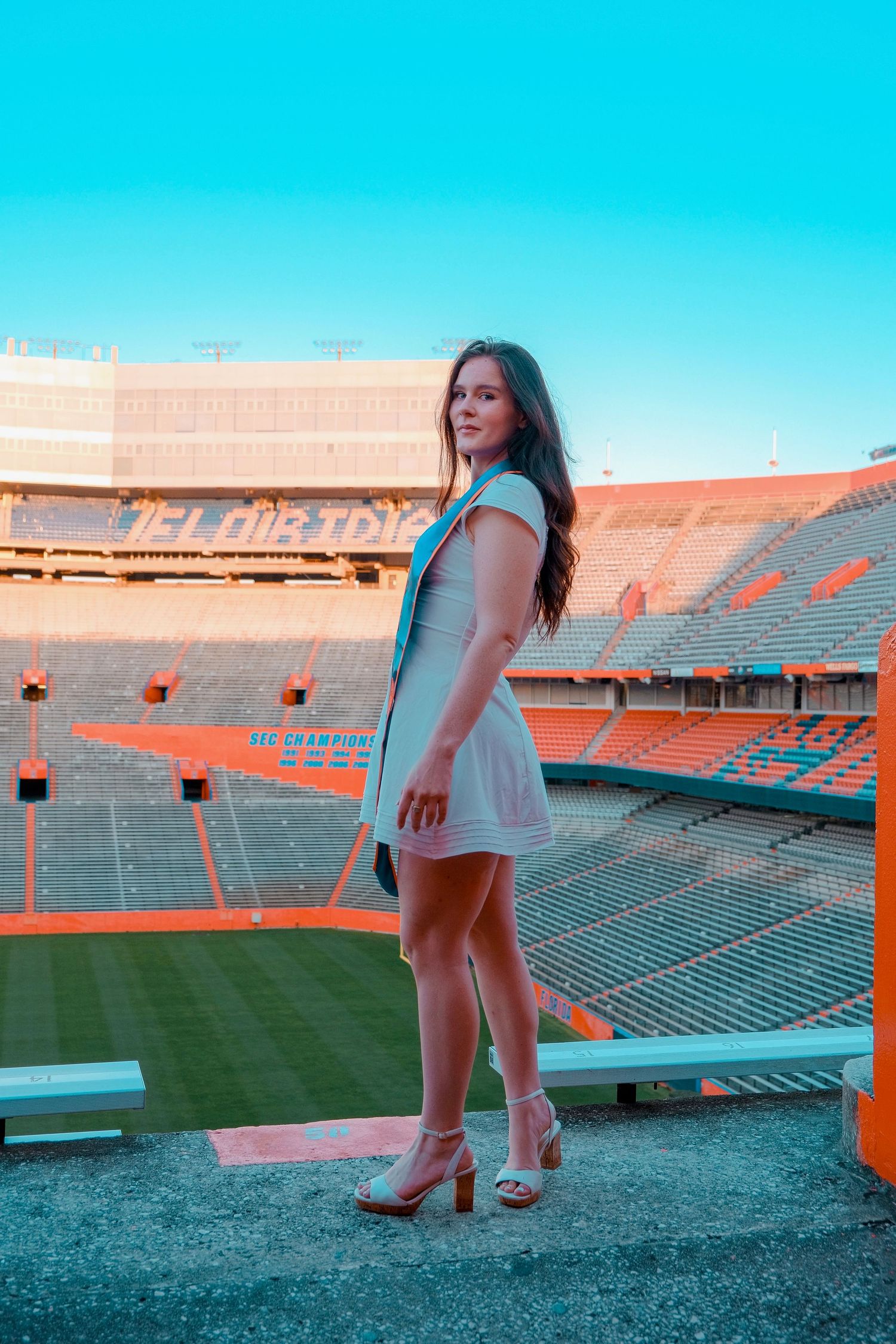 Young woman in white dress and heels posing at an empty orange football stadium on a sunny day.