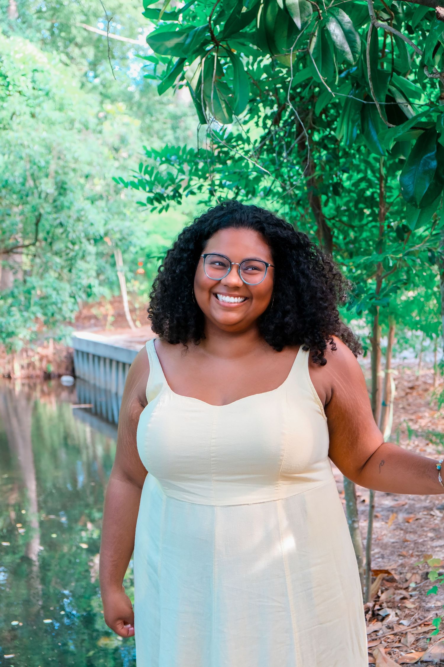 Smiling young woman with curly hair and glasses wearing a light yellow sundress standing outdoors near trees.