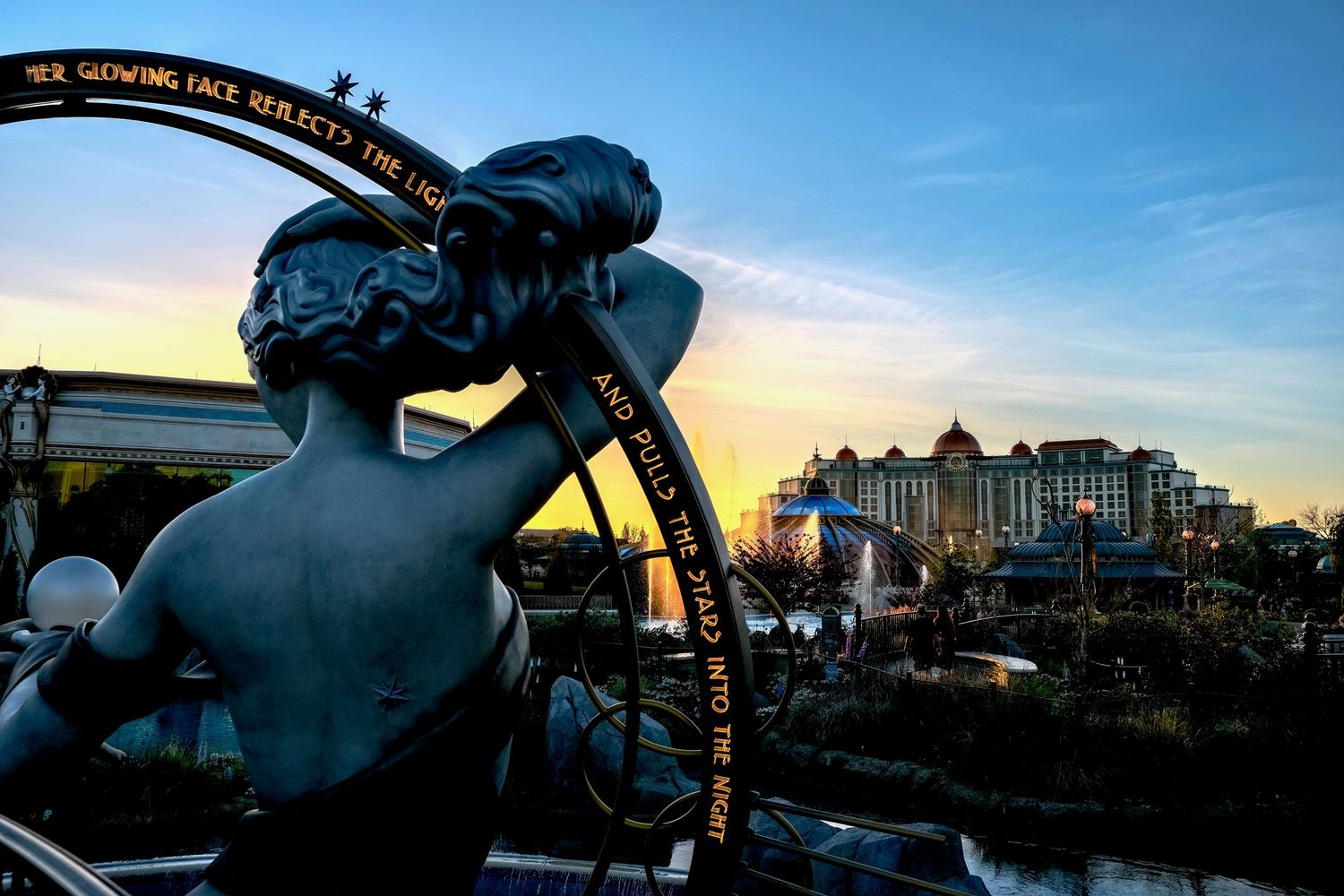 Sunset silhouette of a mermaid statue with golden ring inscription, hotel buildings glowing in background dusk sky.