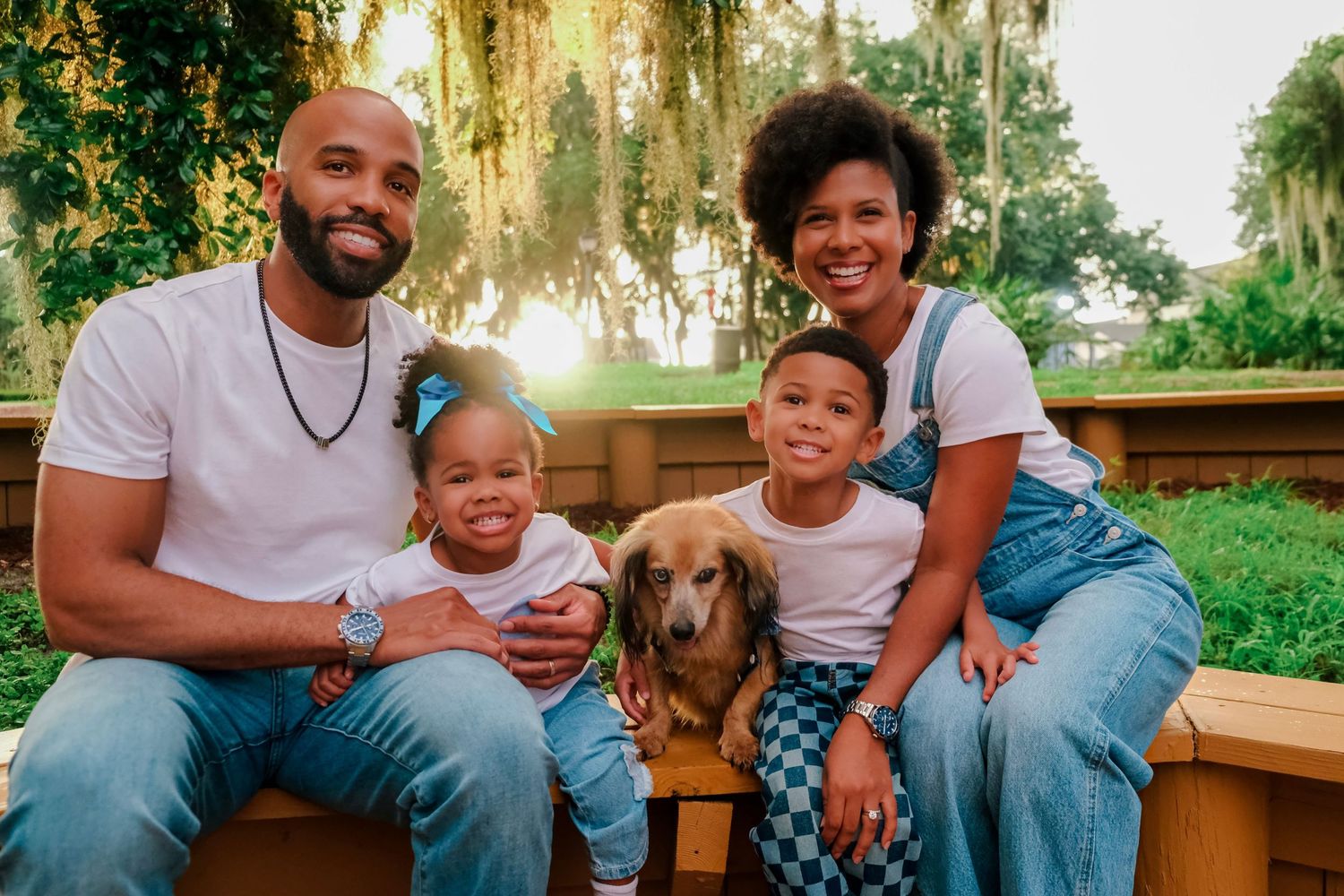 Happy family of four with a golden dog posing outdoors in a lush green park setting during golden hour.