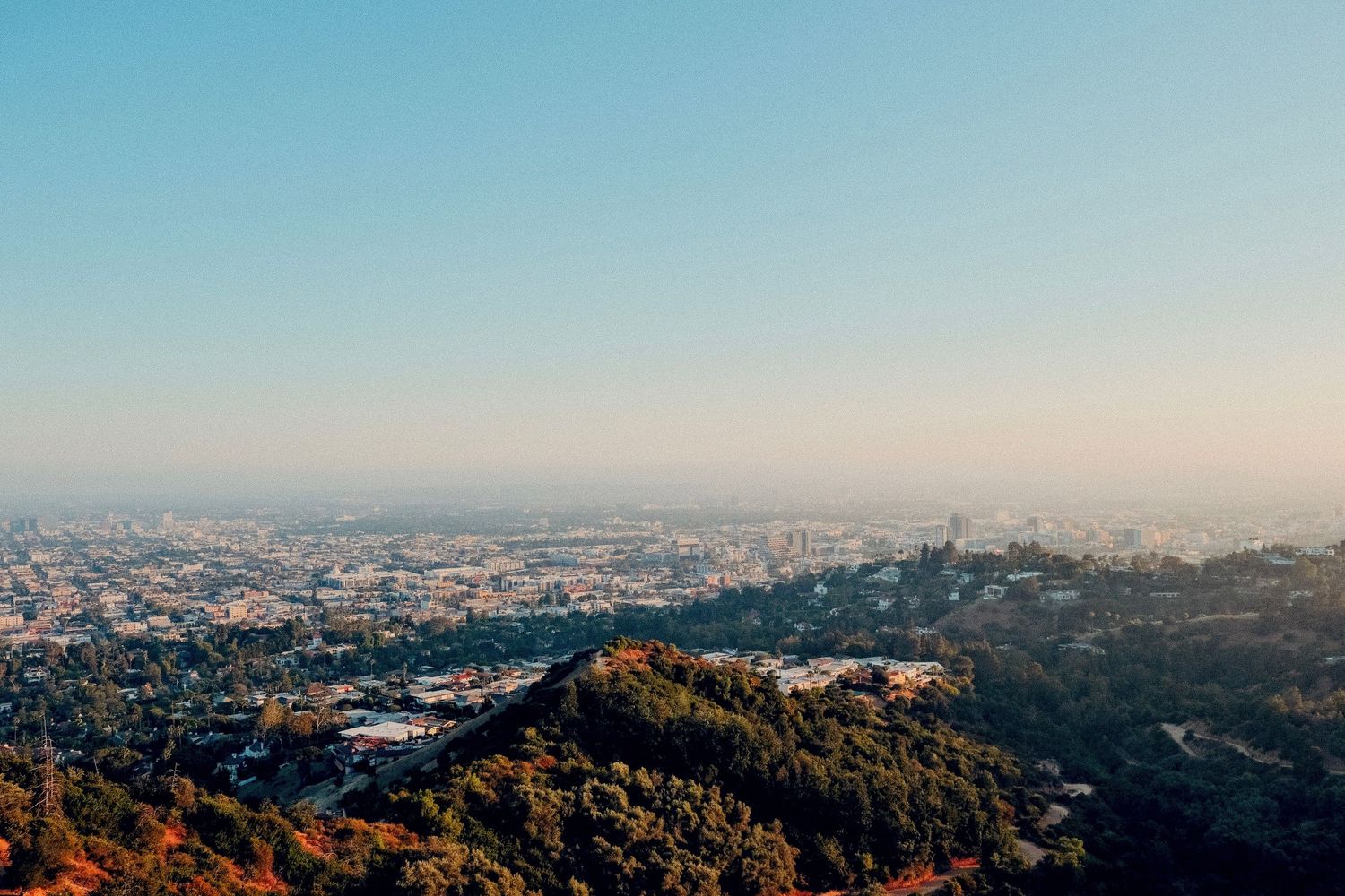 Aerial panoramic view of Los Angeles cityscape with hazy skies, lush green hills, and sprawling urban landscape.
