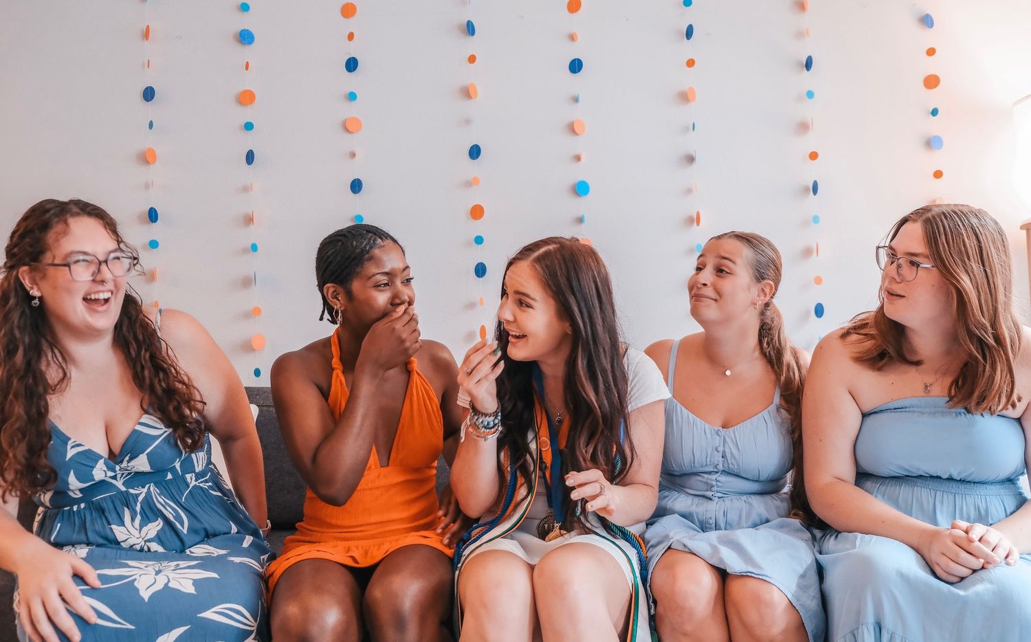 Five young women sitting together on a couch laughing and smiling at a colorful party celebration.