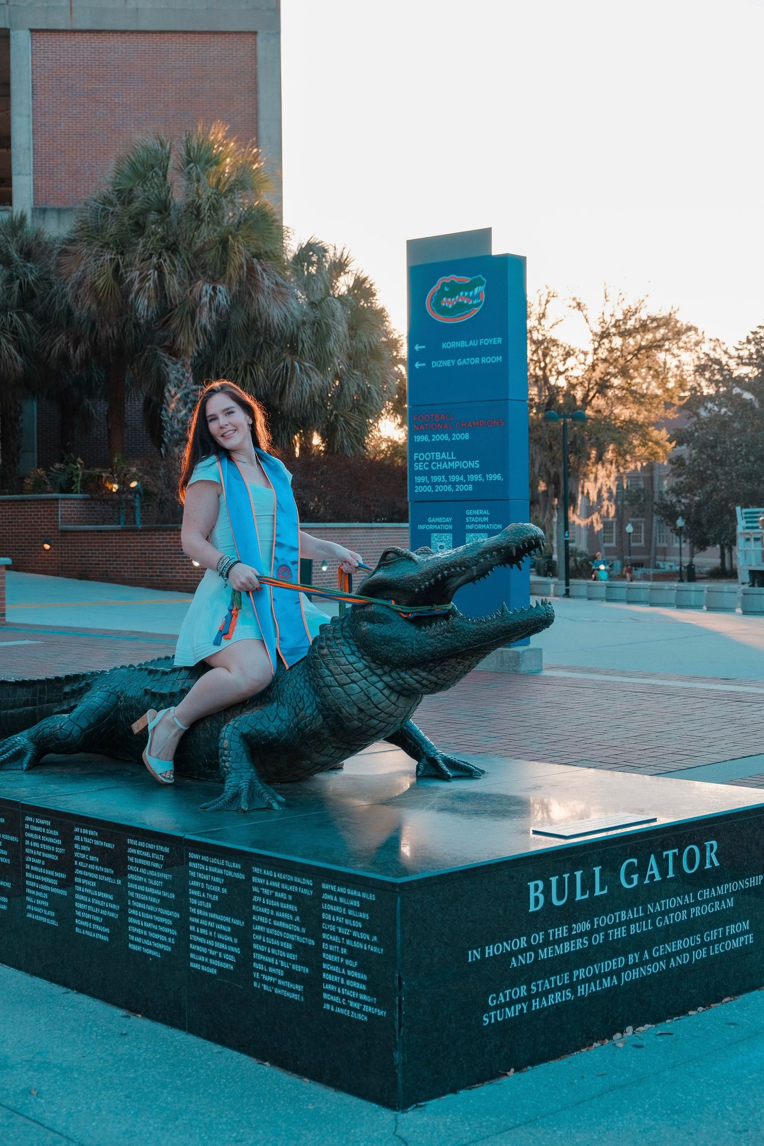 Smiling UF graduate in blue dress poses on Bull Gator statue at University of Florida campus.