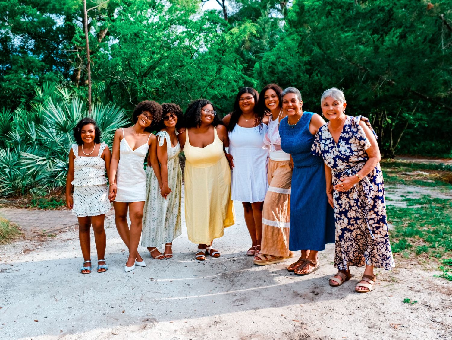 Group of seven women smiling together outdoors on a sandy path surrounded by lush green trees.