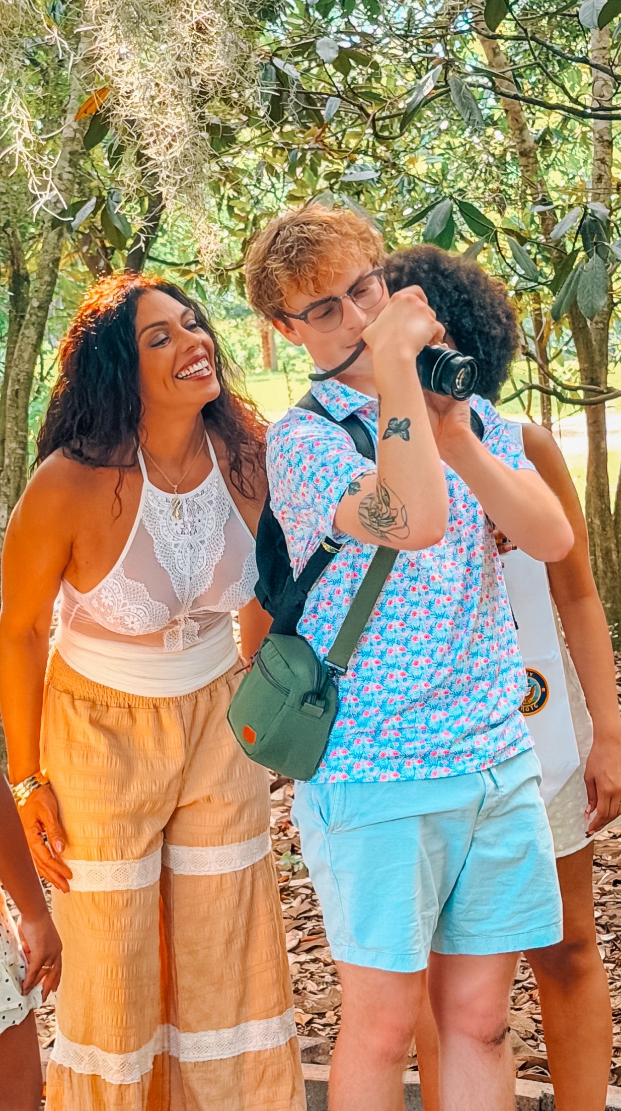 A smiling woman in white top and tan pants stands beside a man photographing with a camera in a sunny outdoor setting.