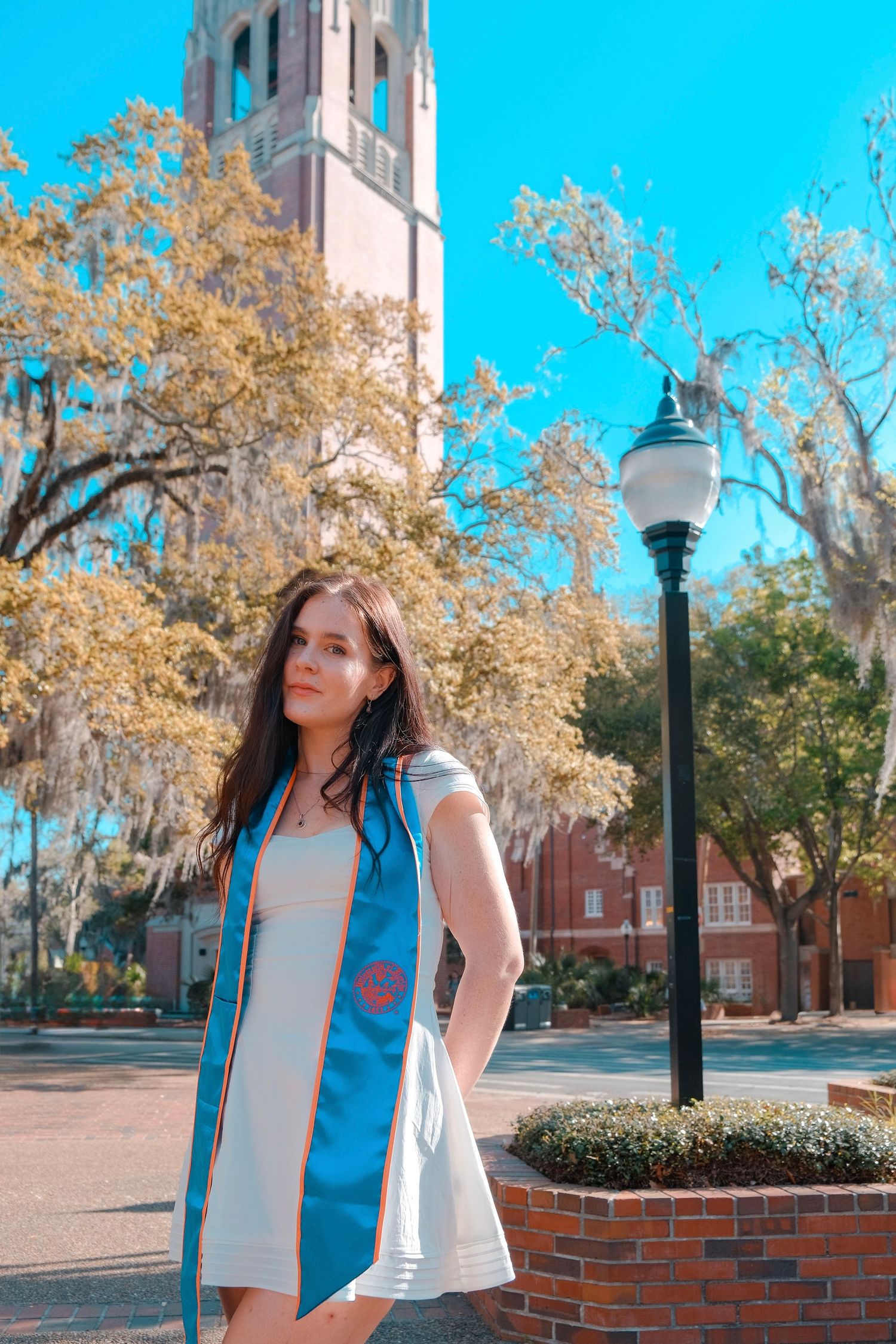 A young woman in a white dress and blue graduation stole poses outdoors near a university tower on a sunny day.