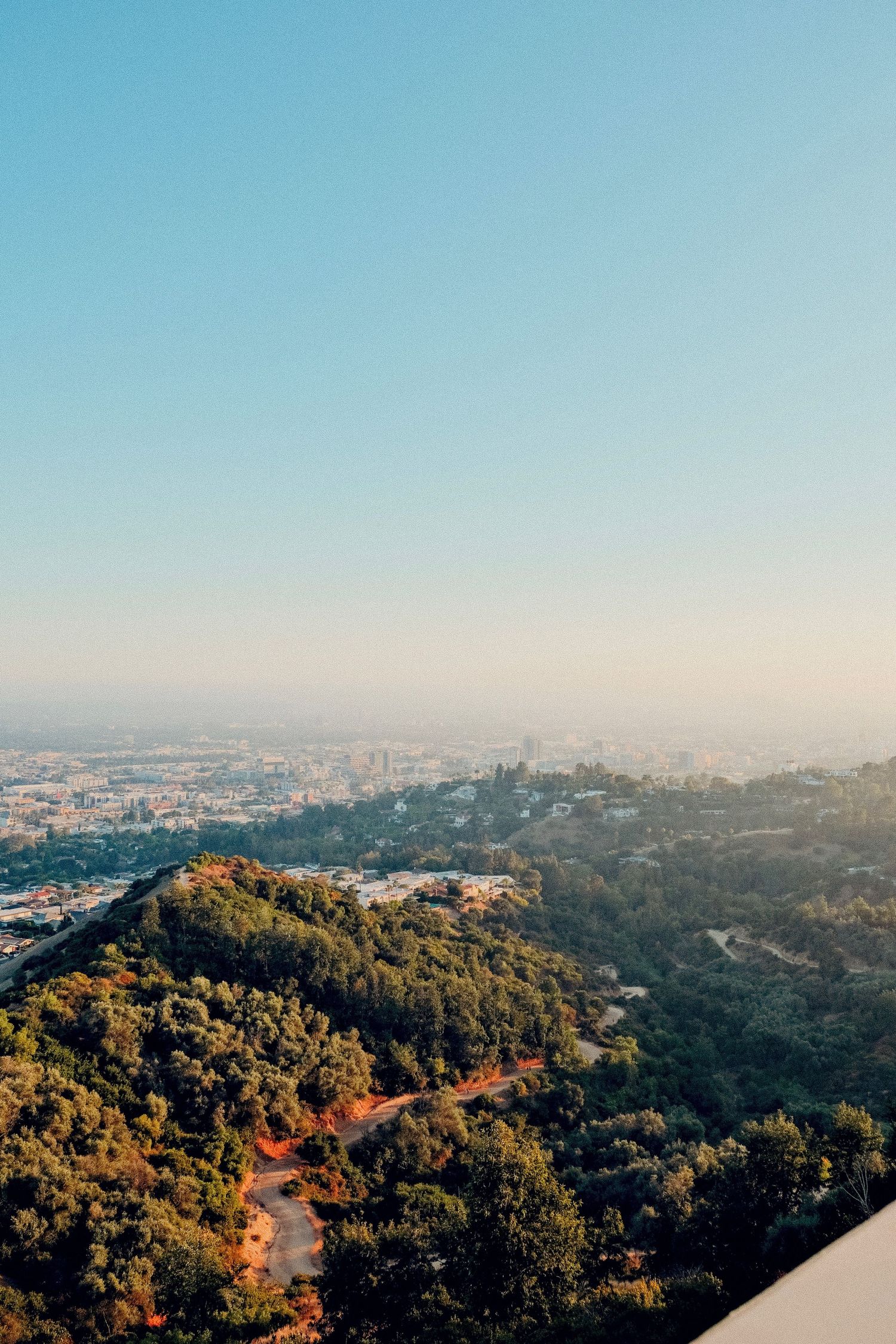 Aerial view of Los Angeles cityscape with lush green hills, winding paths, and hazy skyline at golden hour.