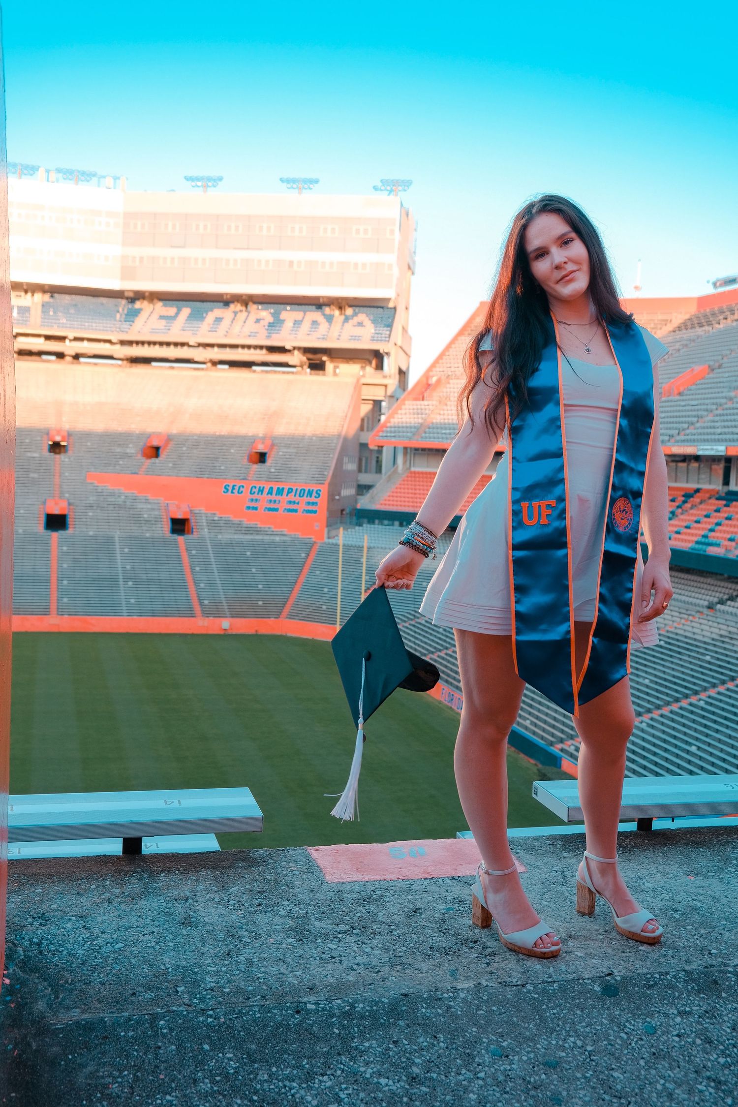Graduate in white dress holds cap with UF stole at Ben Hill Griffin Stadium during golden hour.