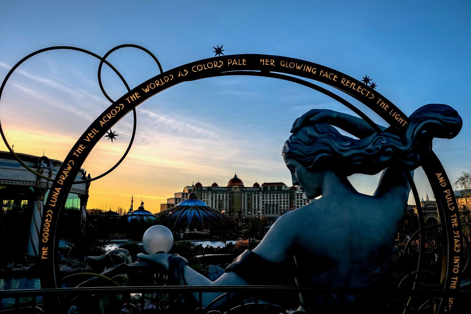 Silhouette of a mermaid statue framed by an ornate ring with text at sunset, with a grand building in the background.