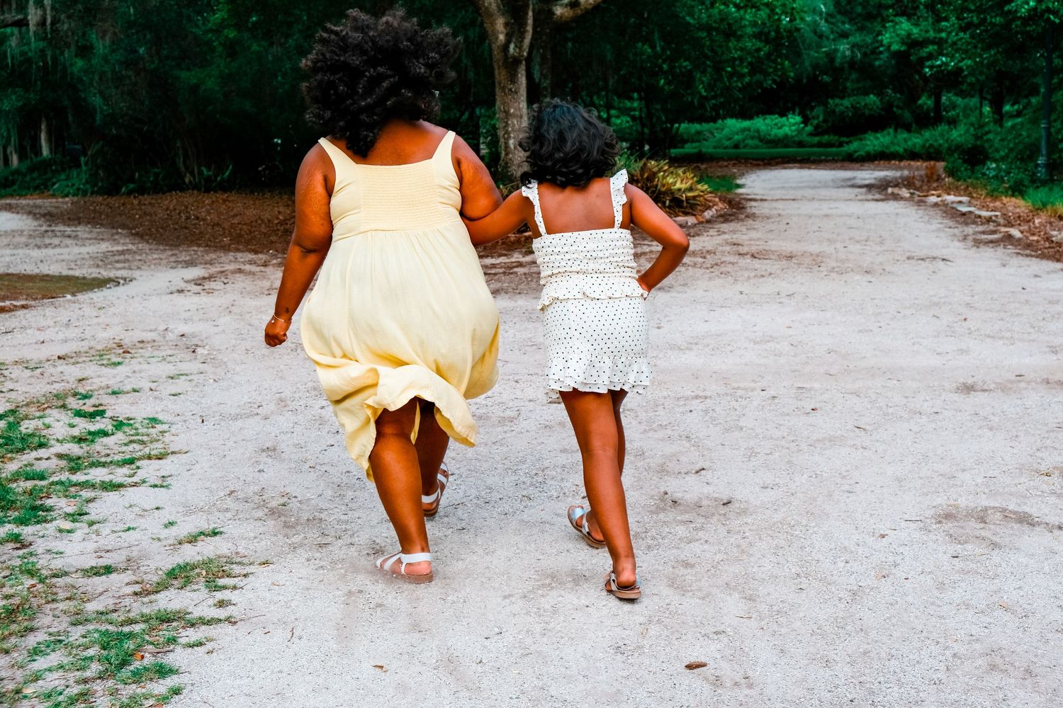 Two Black girls in summer dresses running down a sandy path surrounded by lush green trees.