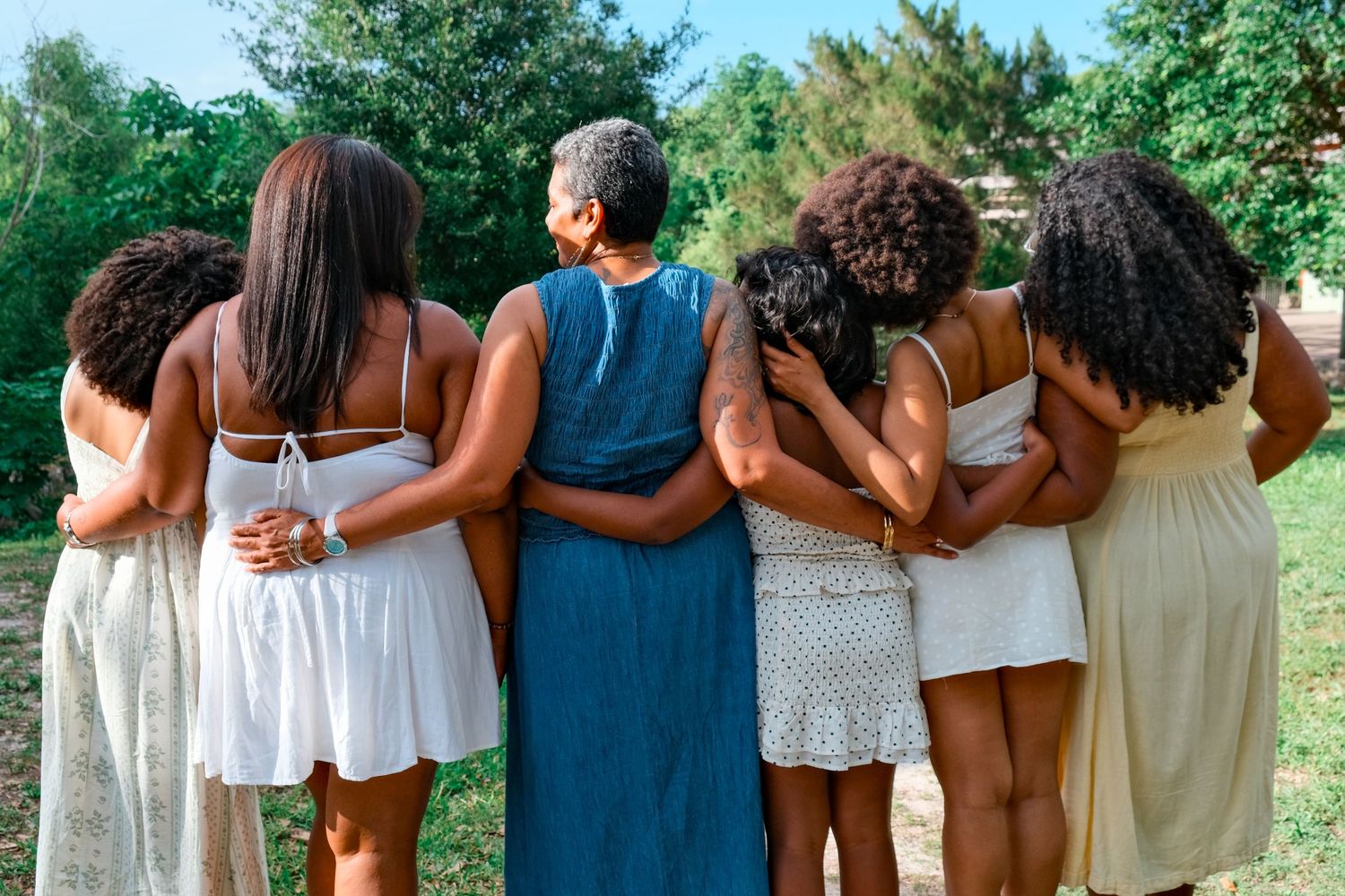 Six women of diverse ages stand arm in arm outdoors, viewed from behind, wearing summer dresses.