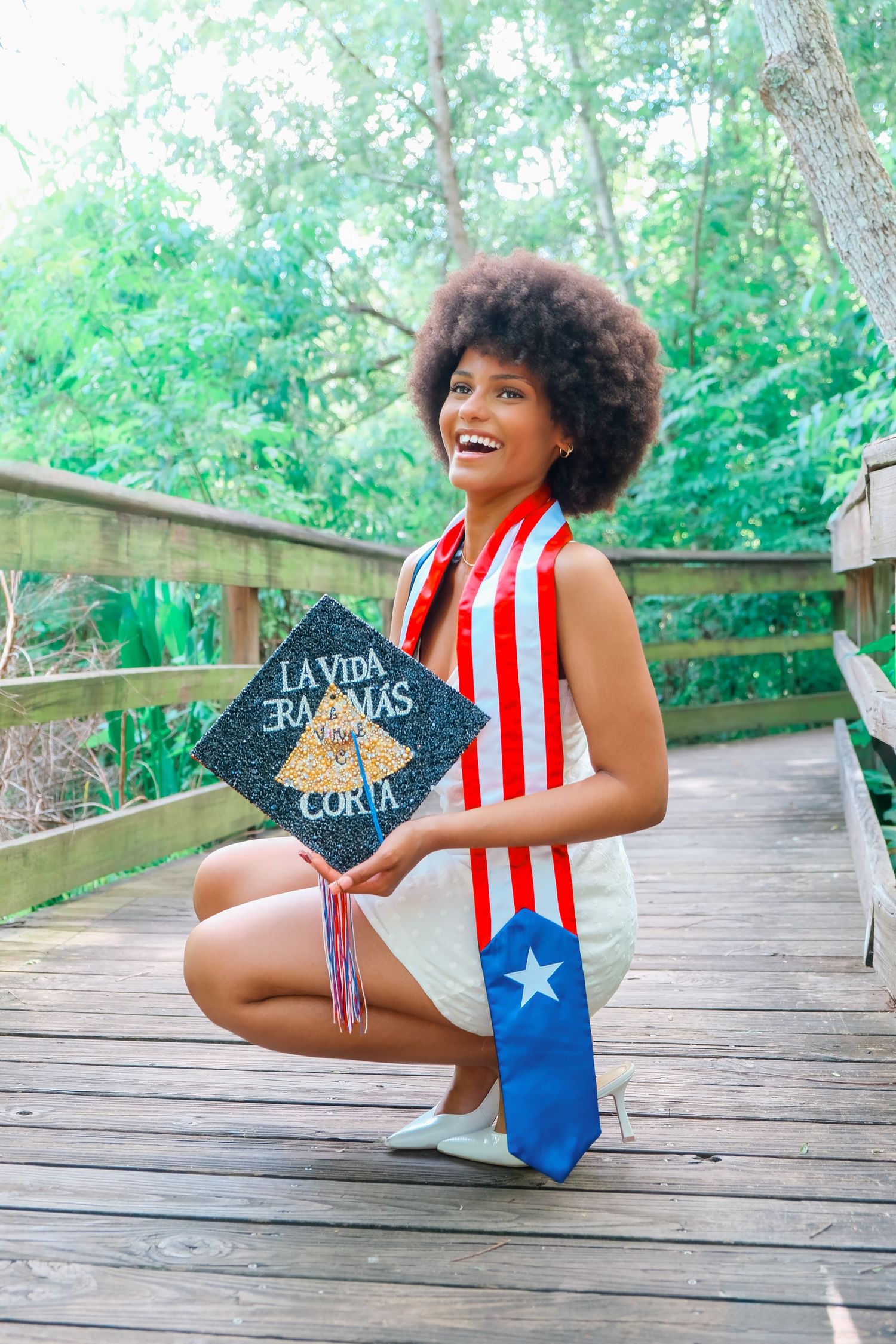 Smiling graduate holding decorated cap reading La Vida Era Mas Corta, wearing Puerto Rican flag sash outdoors.
