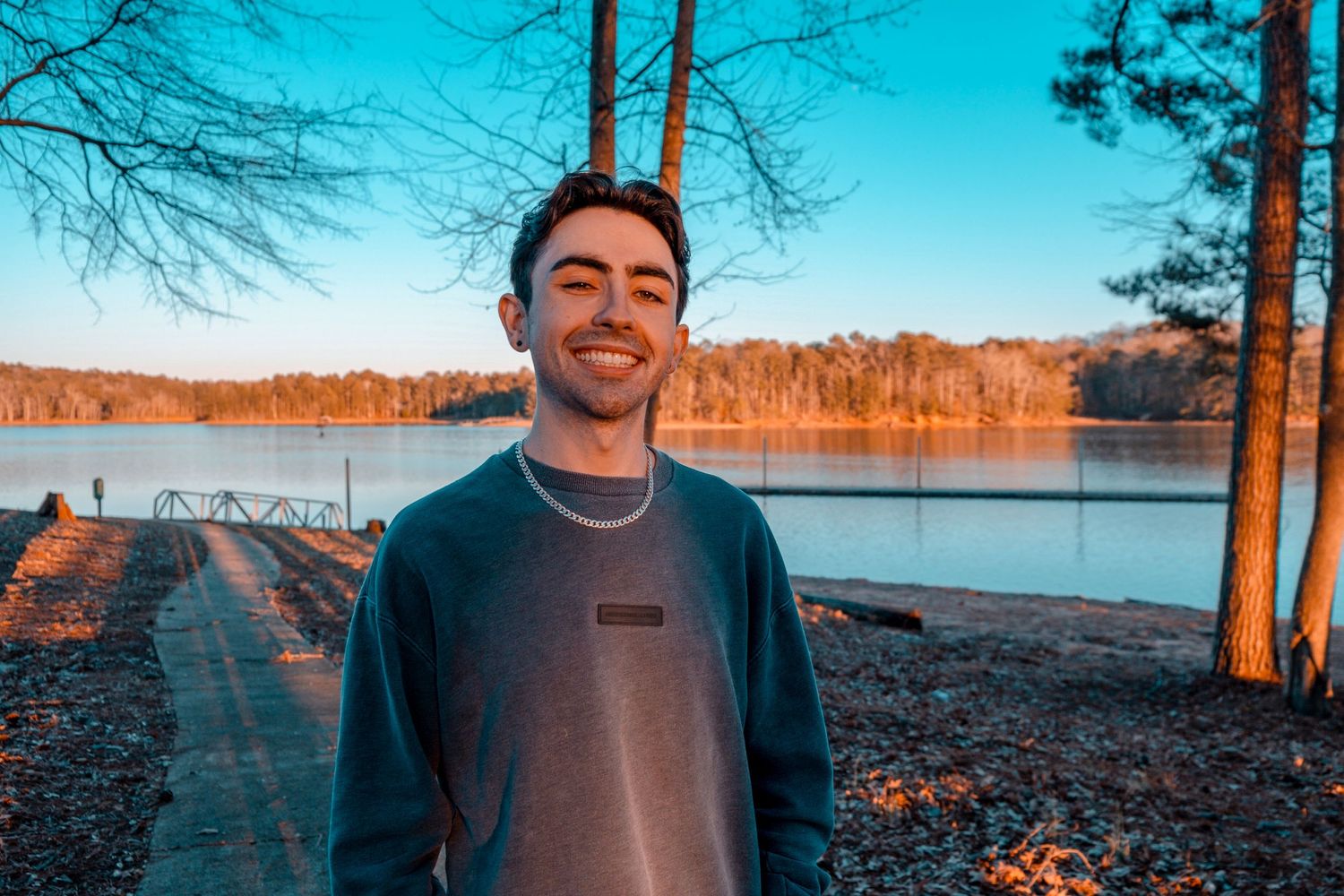 Smiling young man in teal sweatshirt standing near a serene lake with trees and golden hour light.